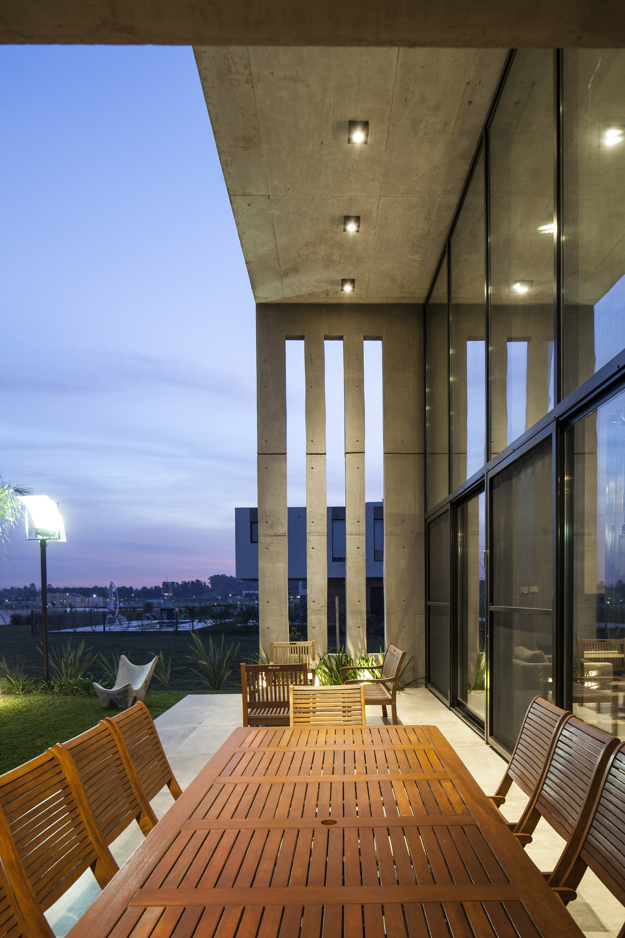Wooden dining table on a covered terrace beside slotted concrete walls and full-height glazing at blue dusk