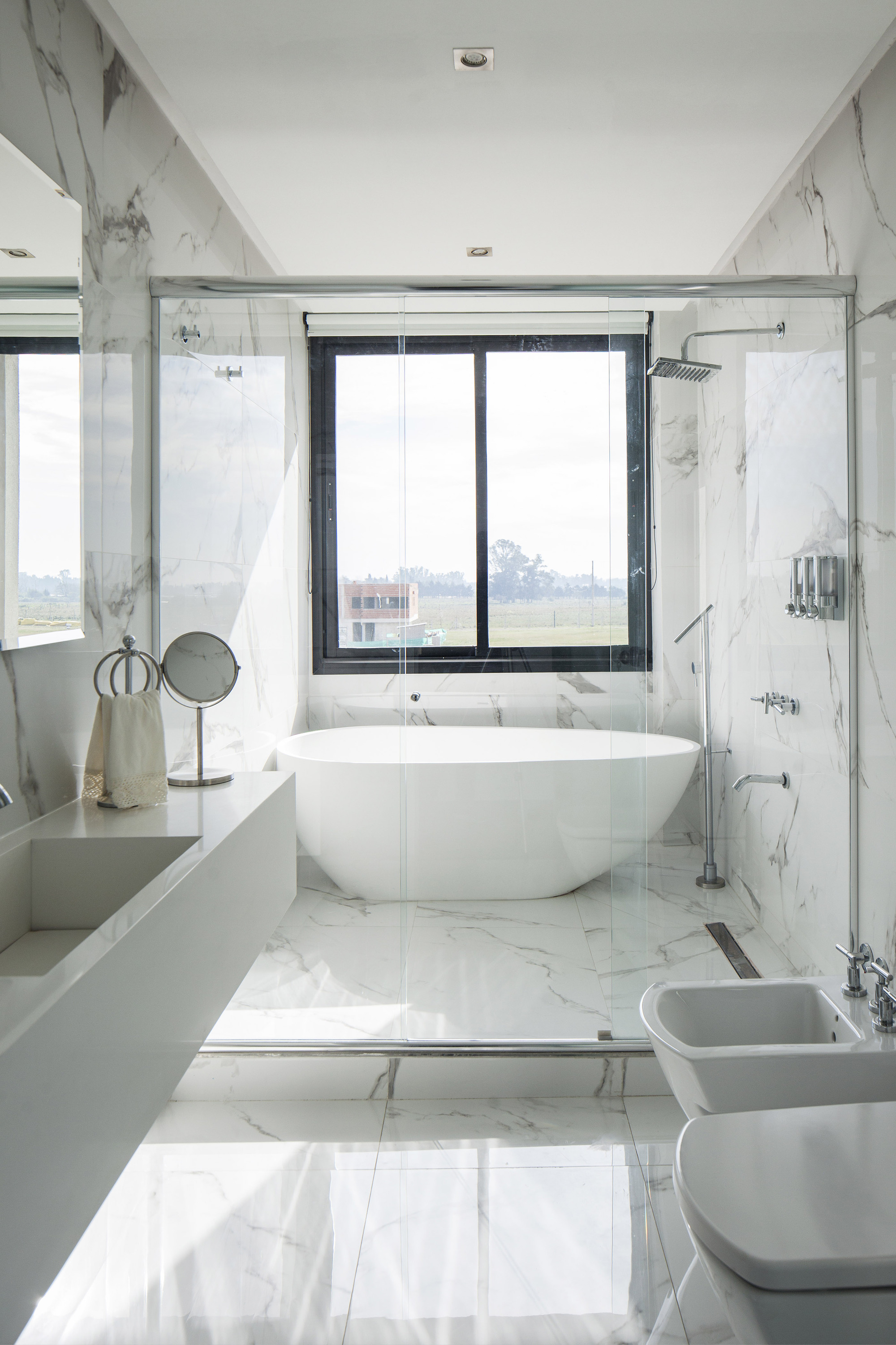 White marble bathroom with a freestanding oval tub behind a glass shower screen and a window onto open fields