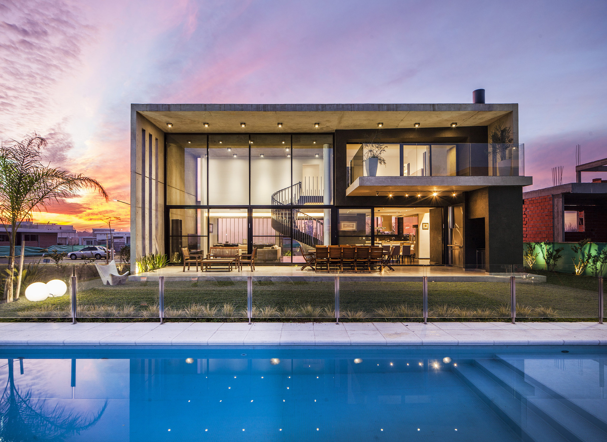 Two-storey concrete and glass house seen across the lit pool and lawn under a vivid orange and violet sunset