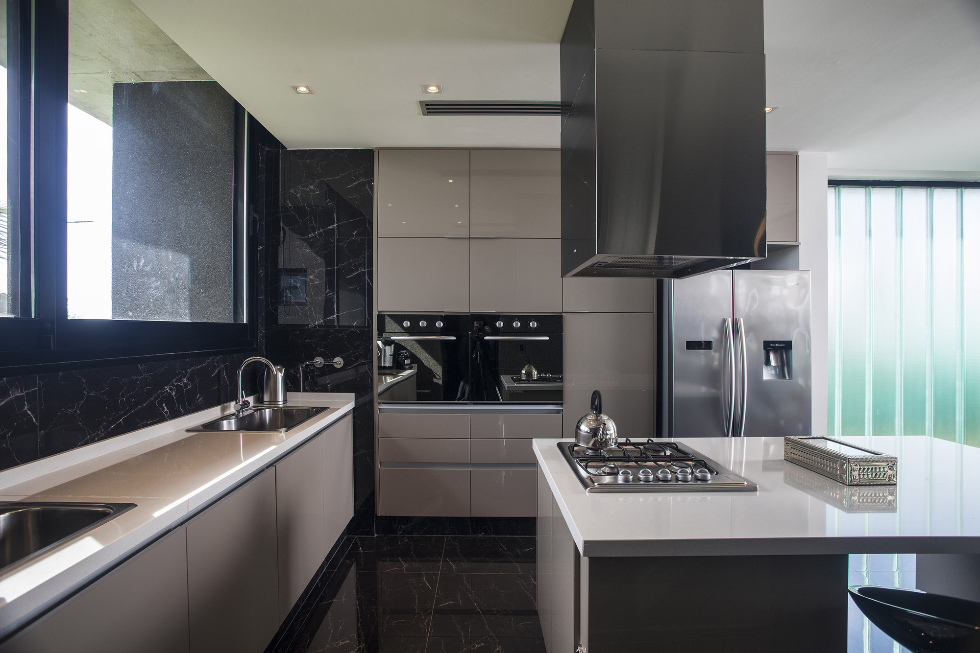 Kitchen with taupe cabinetry, black veined marble backsplash and floor, and a cooktop island lit by daylight from a frosted glass wall