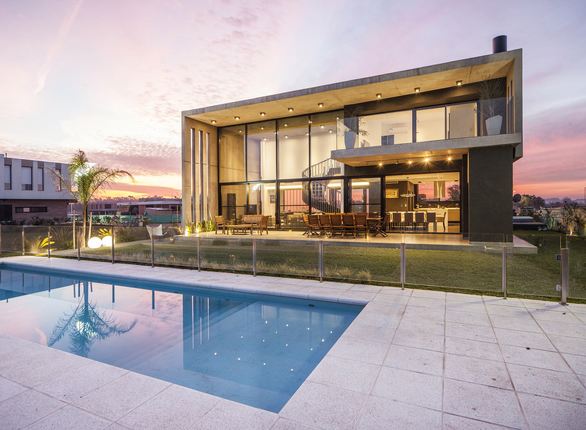 Rear facade of the glazed two-storey house with concrete roof slab, seen across a lit pool at pink dusk