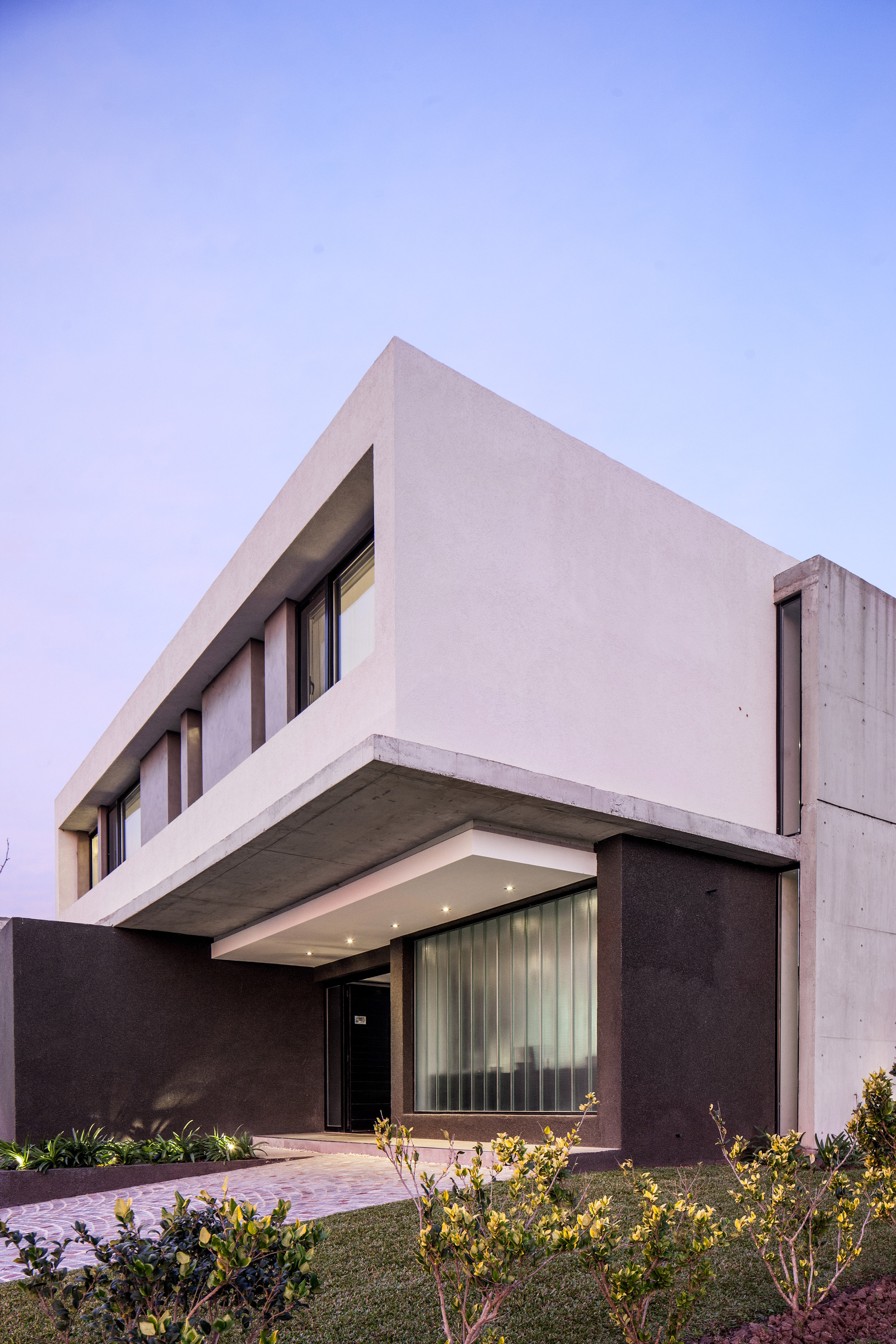 Corner of the white upper volume cantilevering over a dark entrance wall and canopy, with shrubs in the foreground at twilight