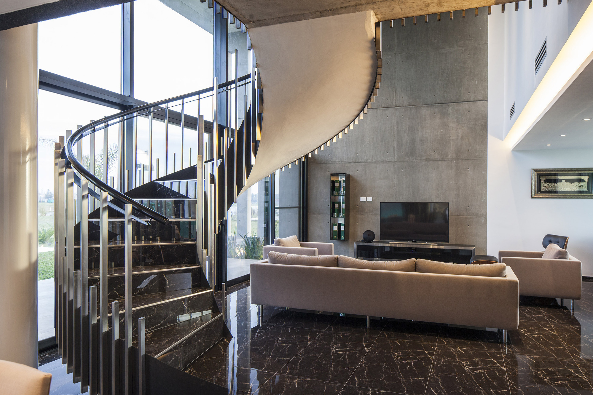 Living room with a curved spiral staircase of steel balusters and black marble treads beside a raw concrete wall in daylight