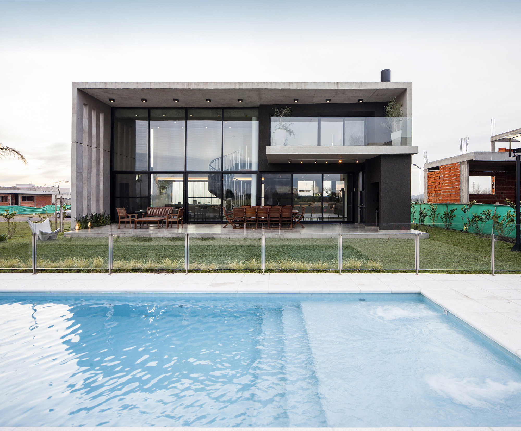 Rear elevation of the concrete and glass house behind a rippling pool, with an unfinished brick building beside it under overcast sky