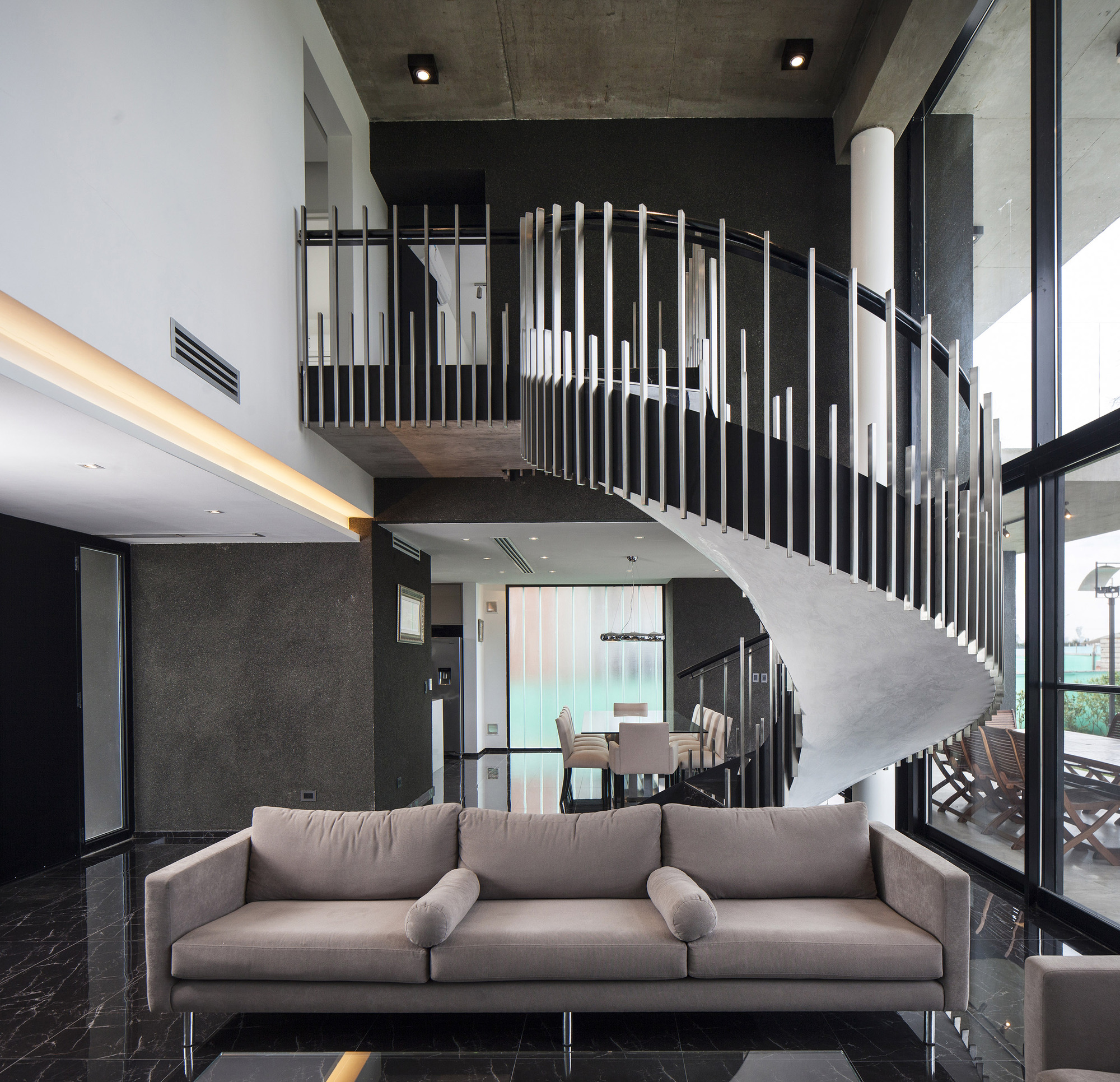 Spiral stair with slender steel balusters rising above a taupe sofa and black marble floor, with garden light through the glazing