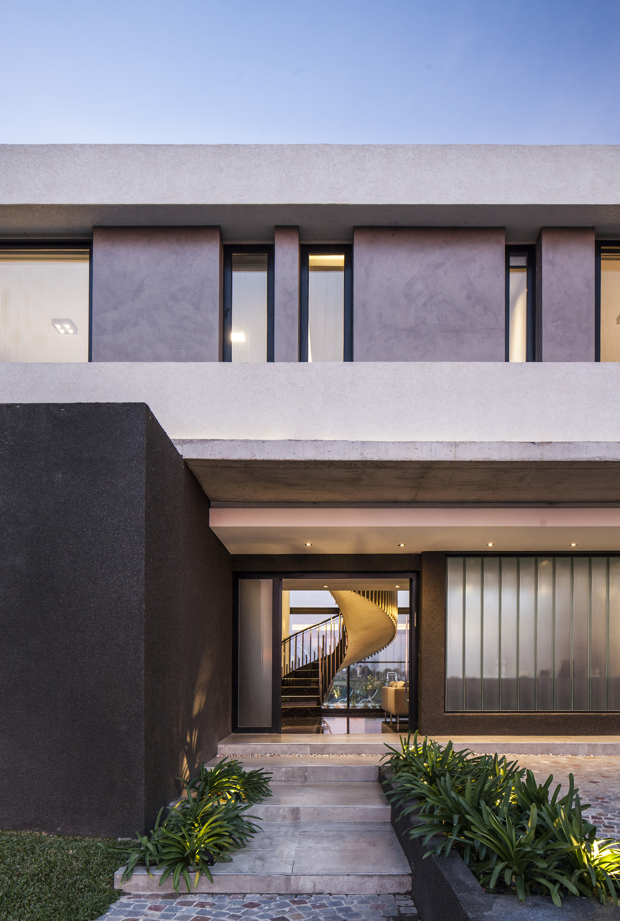 Entrance facade with a glazed door framing a spiral staircase, lit planting beds and stone steps at dusk