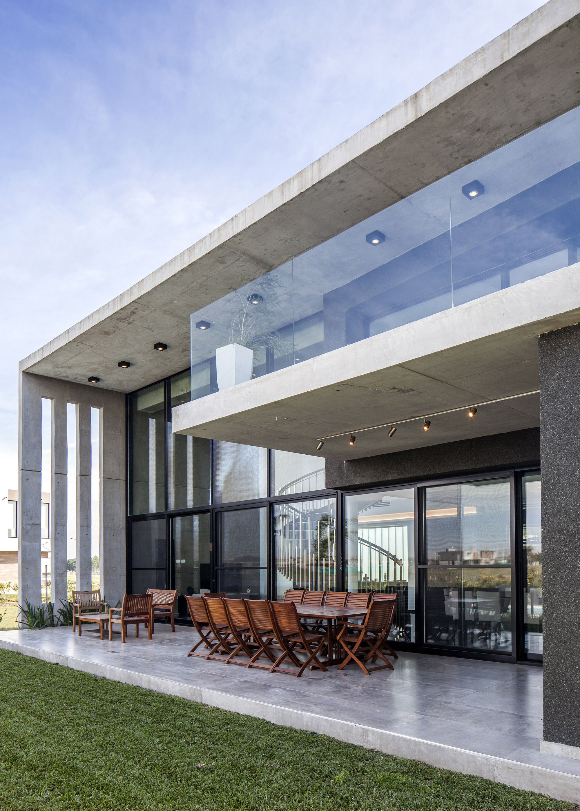 Covered terrace with a timber dining table beneath a concrete slab and glass balustrade, on a lawn in daylight
