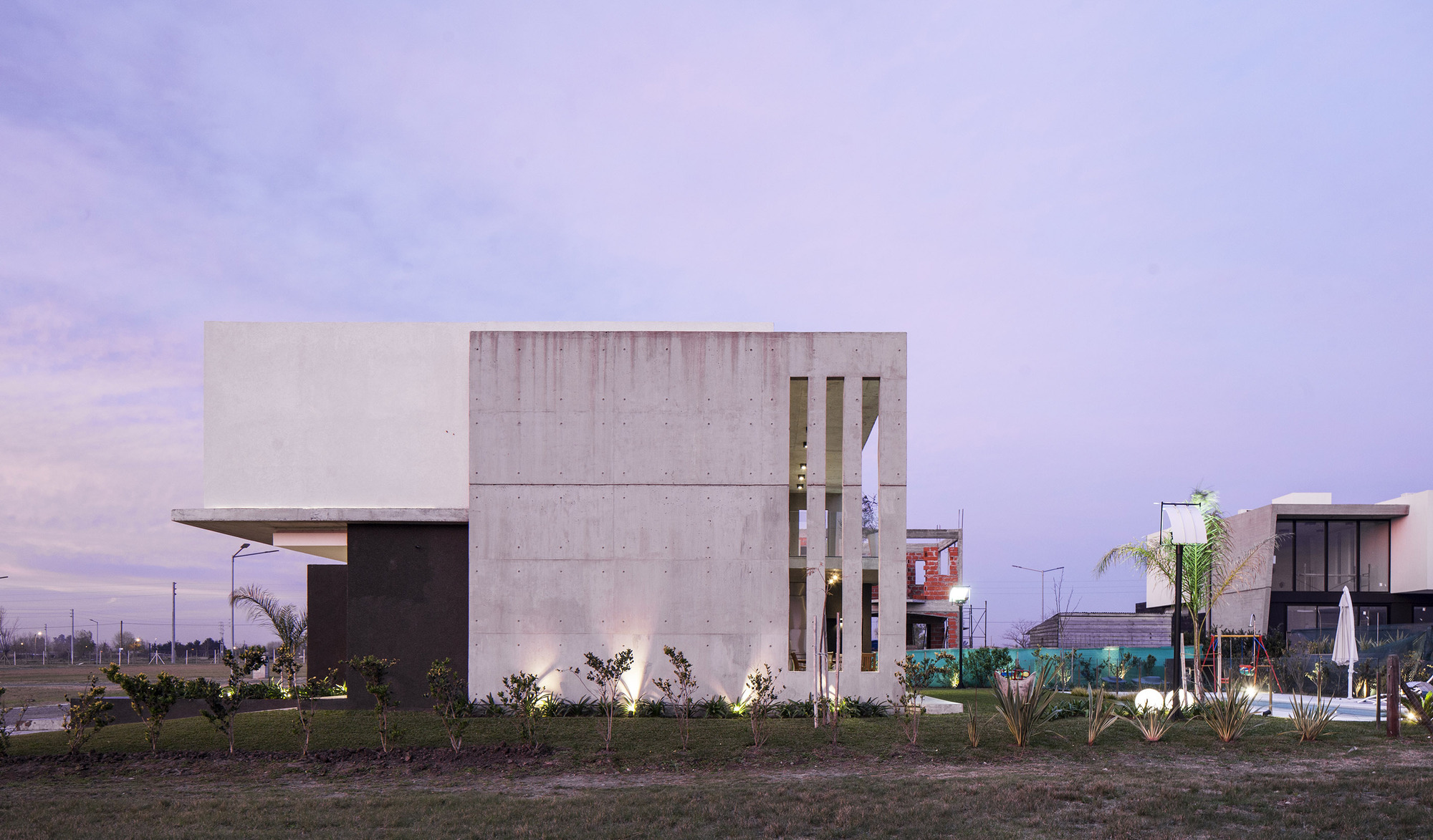 Side elevation of the board-marked concrete volume with a slotted opening and uplit shrubs under a violet twilight sky