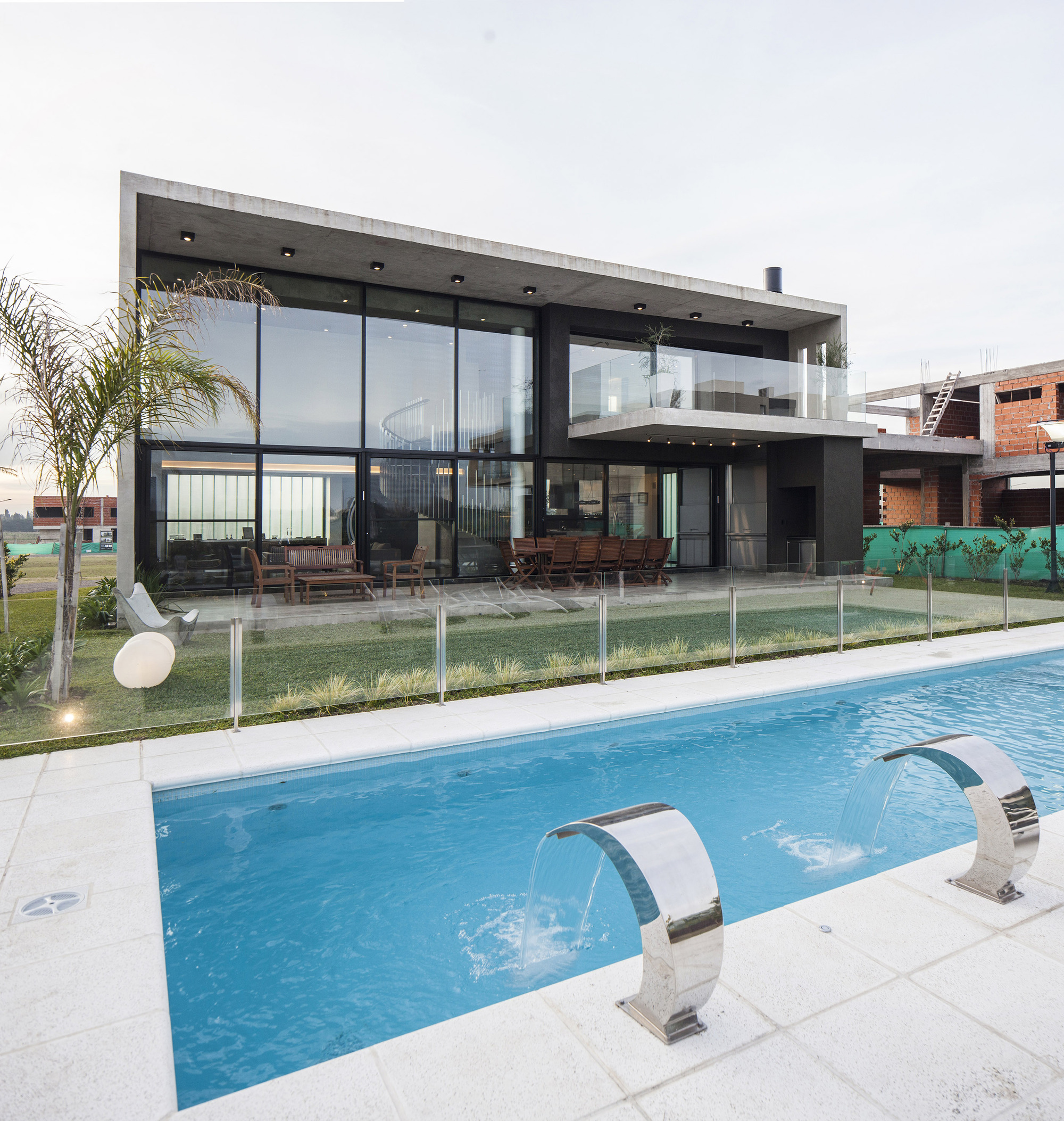 Rear facade seen across the pool with steel water spouts, glass walls and a cantilevered balcony under overcast light