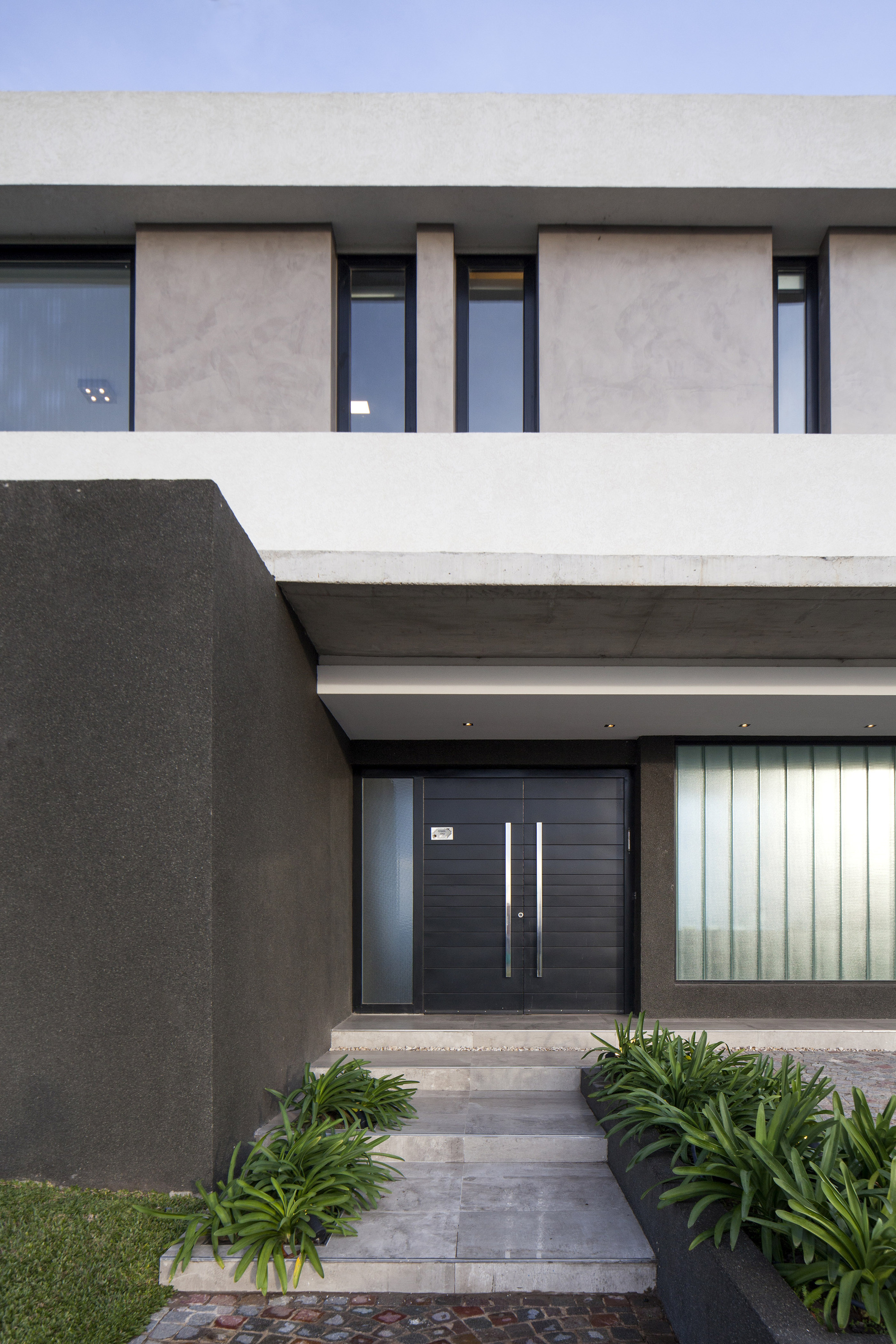 Entrance with a black double door, ribbed translucent glass panel and stone steps flanked by green plants in daylight