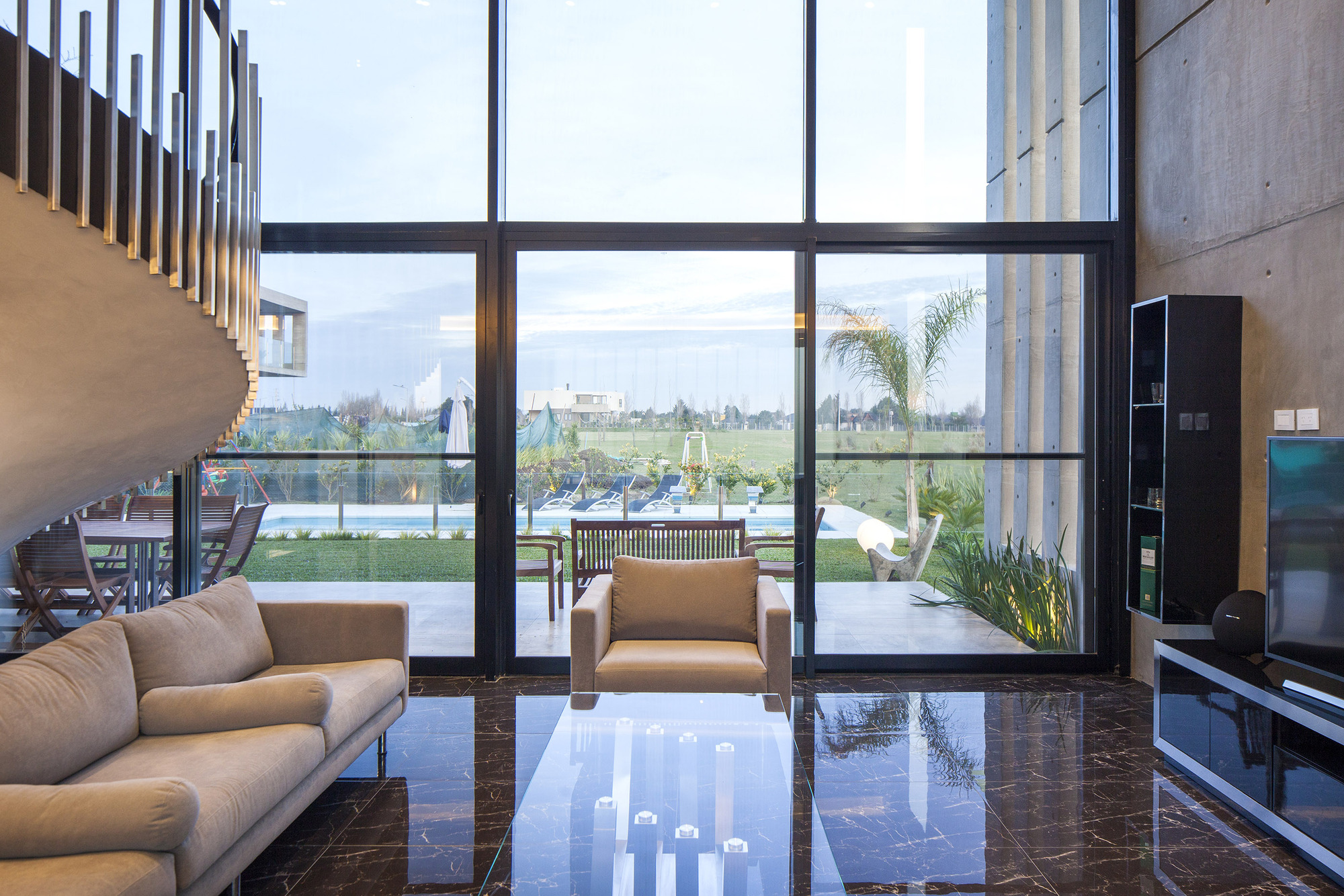 Living room with a curved stair balustrade, tan sofa and armchair facing floor-to-ceiling glass that opens to the lawn and pool
