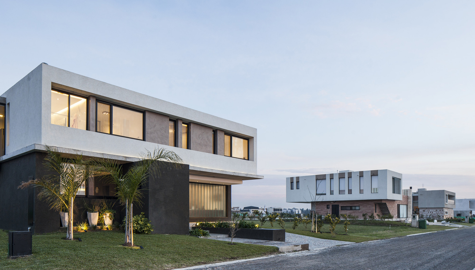 Two-storey white concrete volume with a dark stone base and palms in the foreground, with a neighbouring house across the lawn at dusk