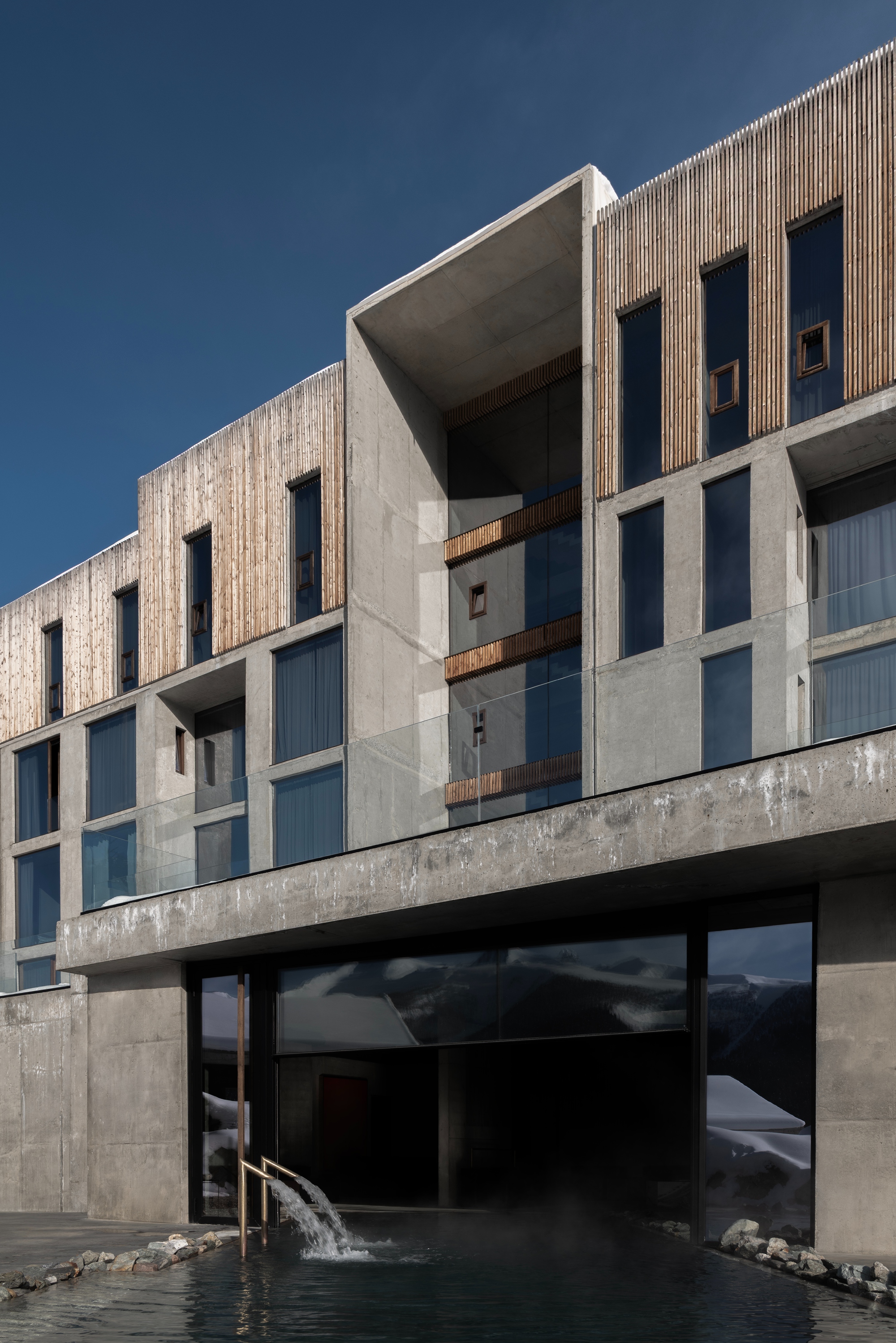 Elevated concrete structure with vertical timber fins above stone-edged water feature and hillside backdrop