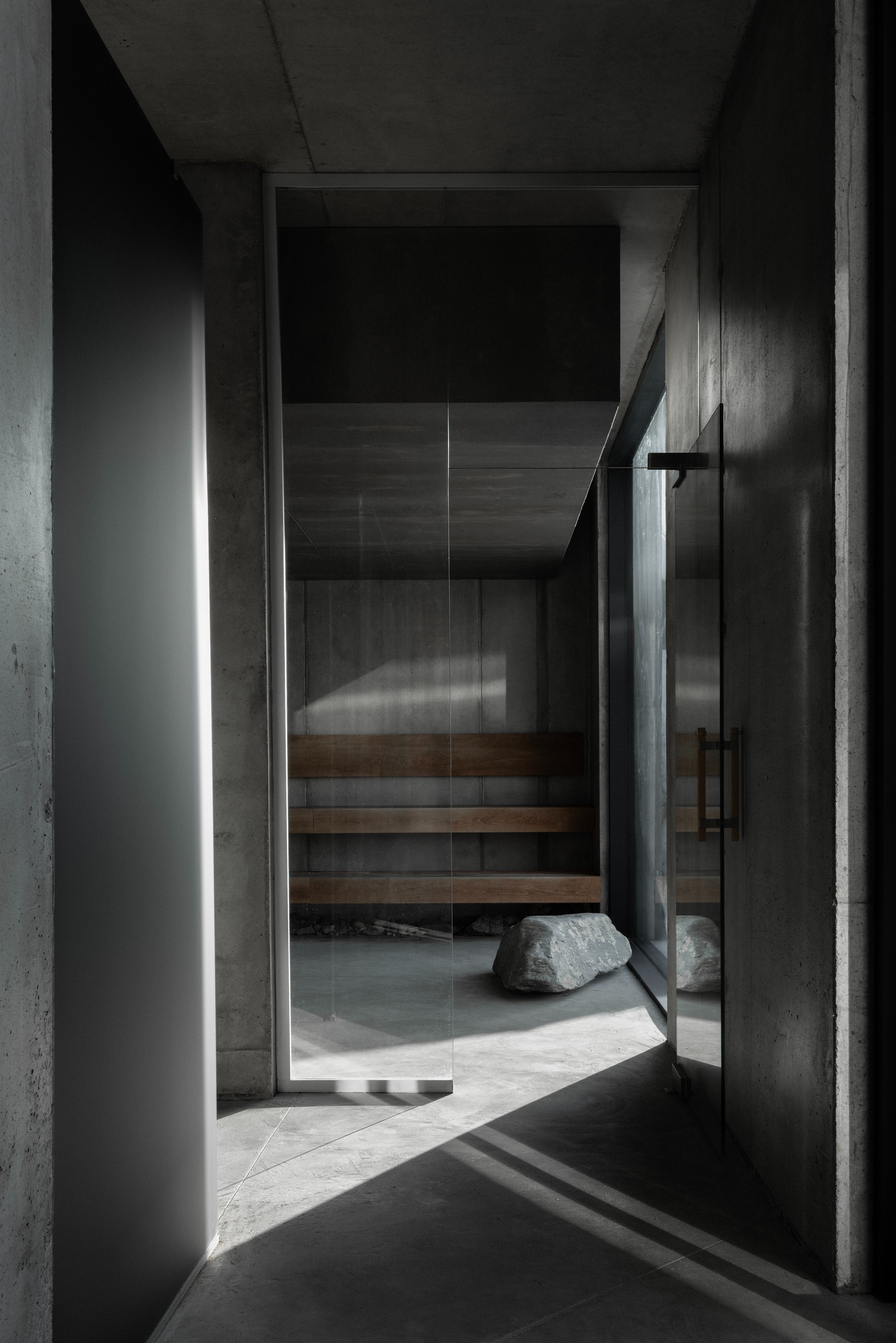 Glass door entry to a timber sauna with a boulder on concrete floor illuminated by vertical light slot