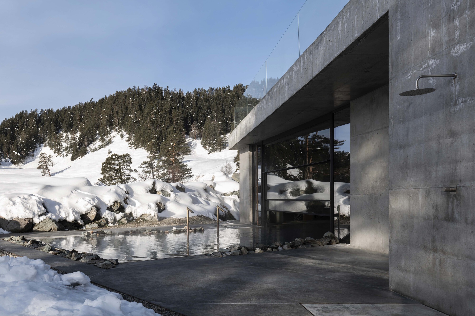 Concrete pavilion with glass walls and reflecting pool beside a snow-covered hillside with evergreens