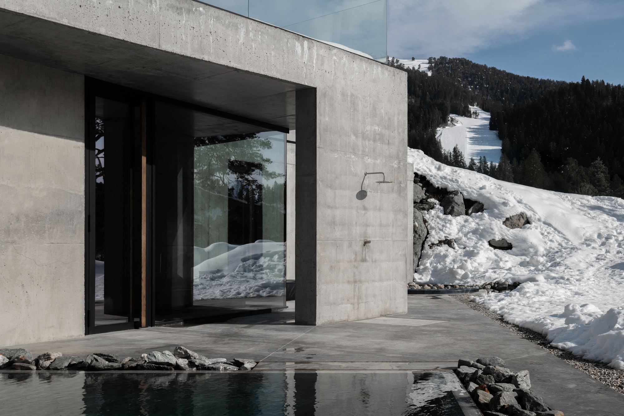 Outdoor shower fixture mounted on concrete wall next to reflecting pool and snowy mountain slope