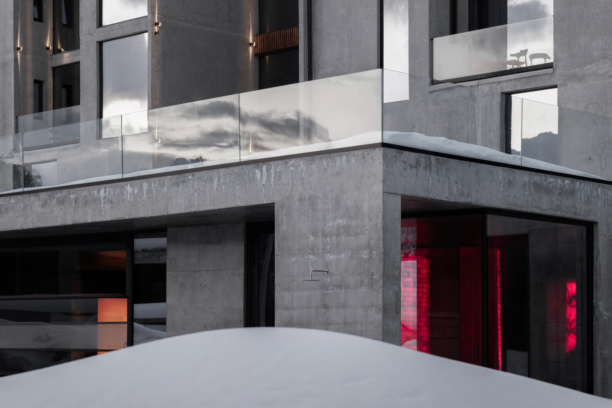 Concrete courtyard with glass balustrades and red-lit interior spaces visible through ground-floor openings in snow