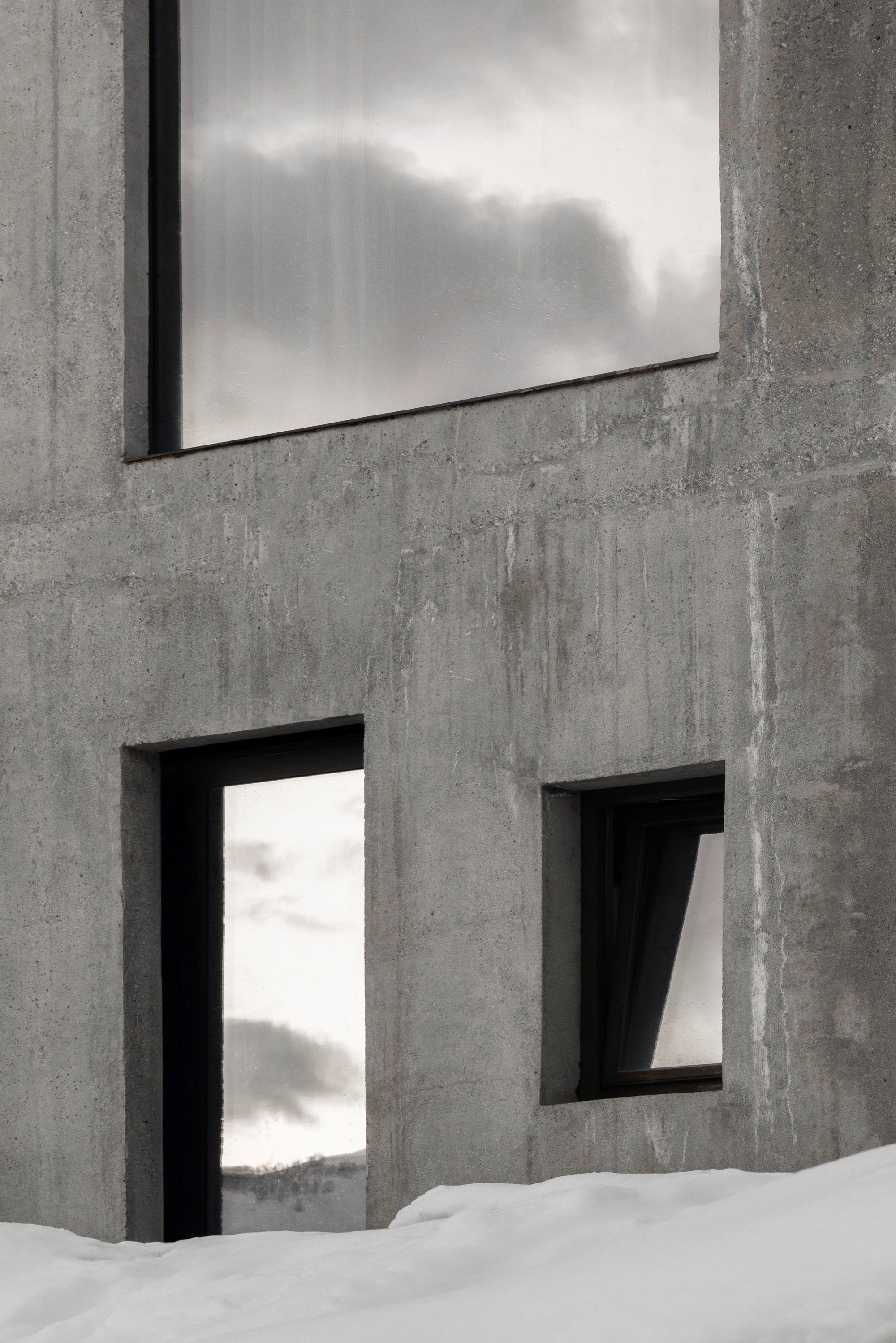 Board-formed concrete facade with rectangular window openings and deep reveals beside snowdrift