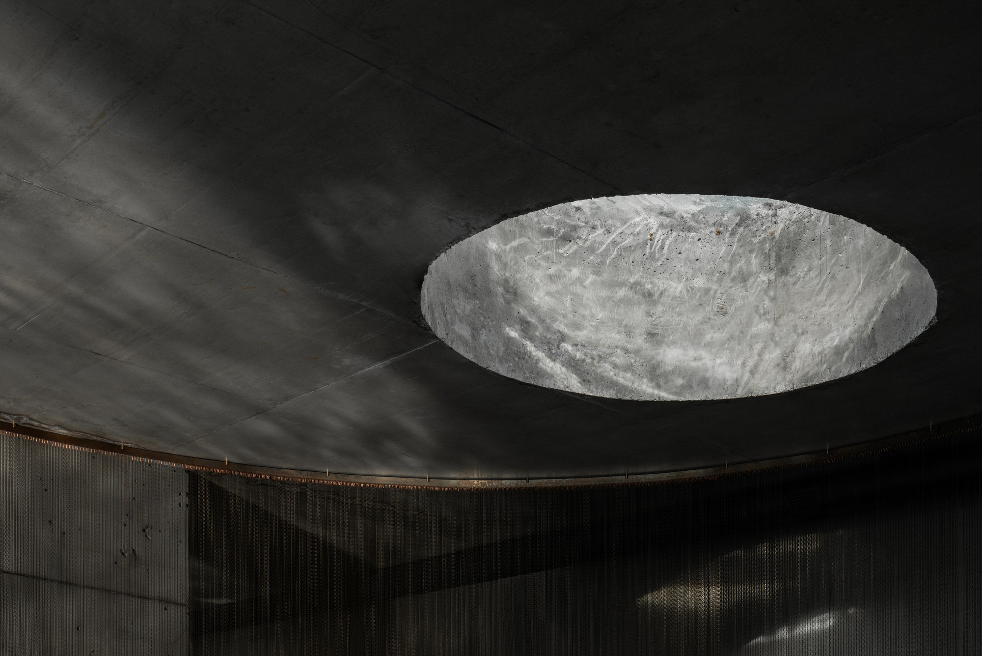 Circular skylight cut into blackened concrete ceiling creating a pool of daylight below