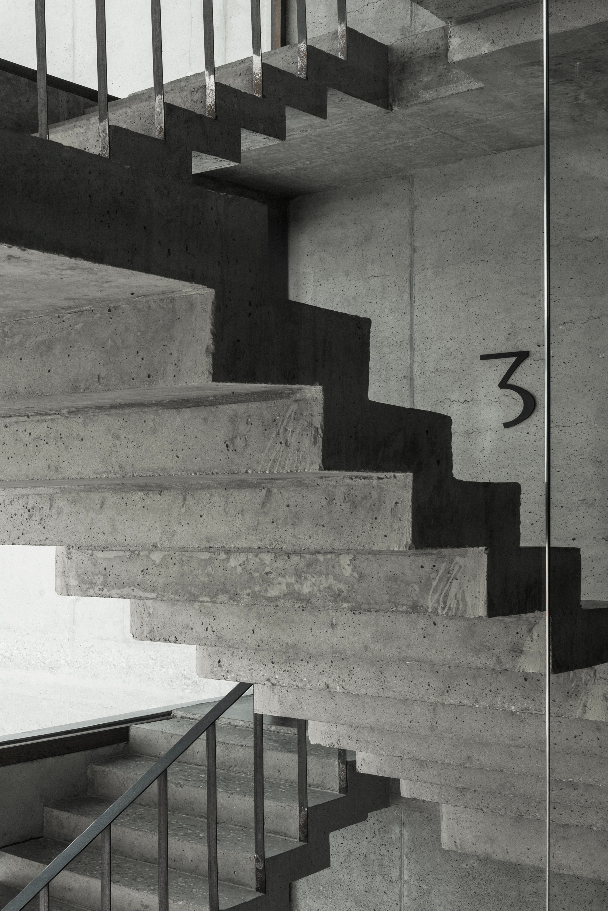 Folded concrete staircase with exposed formwork texture and steel handrail under natural light from a skylight