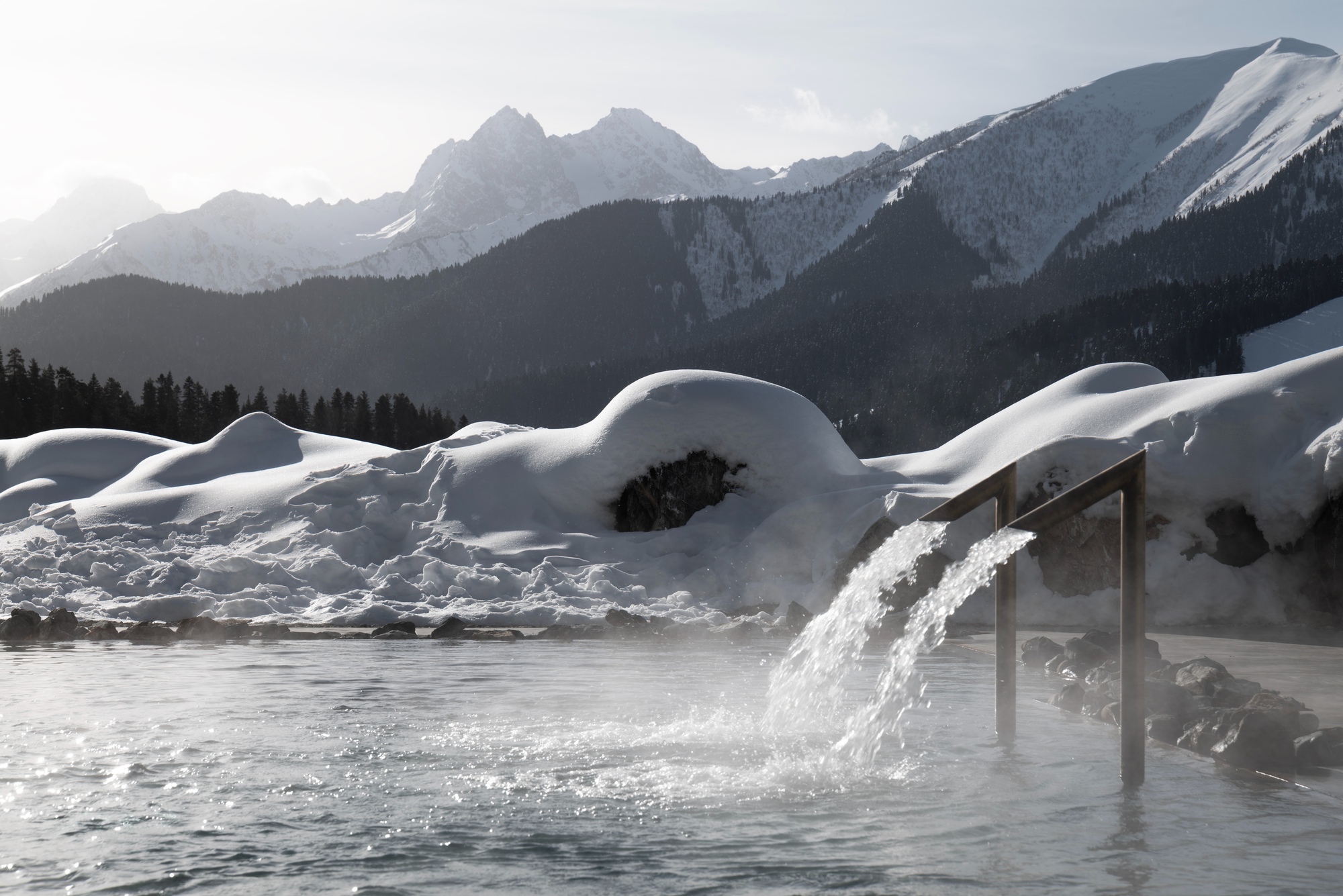 Outdoor pool with cascading water feature set against snow-covered landscape and mountain peaks at dusk