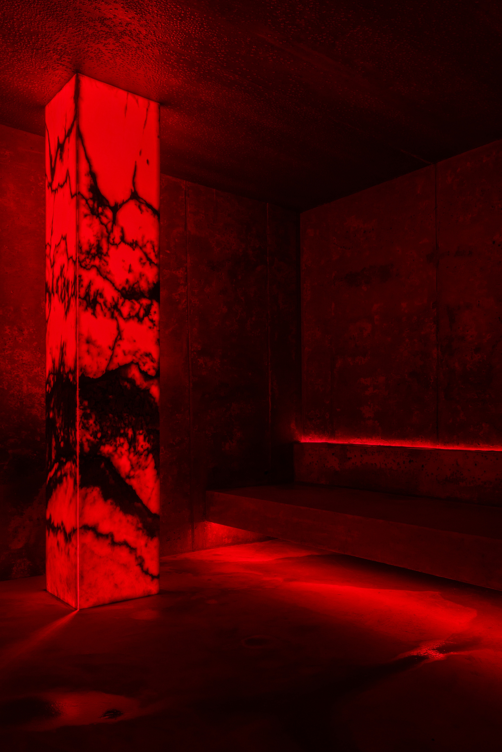 Red-lit spa room with translucent illuminated column and low horizontal bench at night