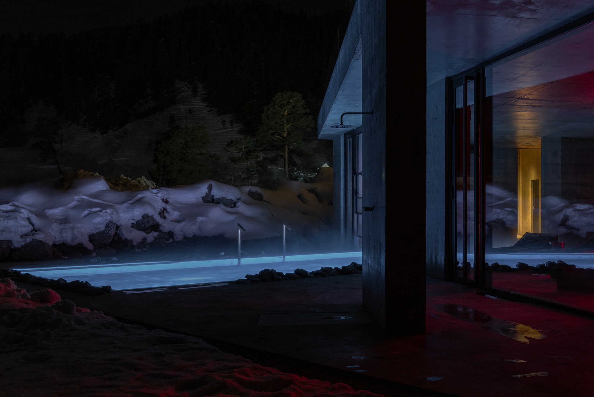 Illuminated outdoor pool at night viewed from interior through concrete columns with snowy rock formations beyond