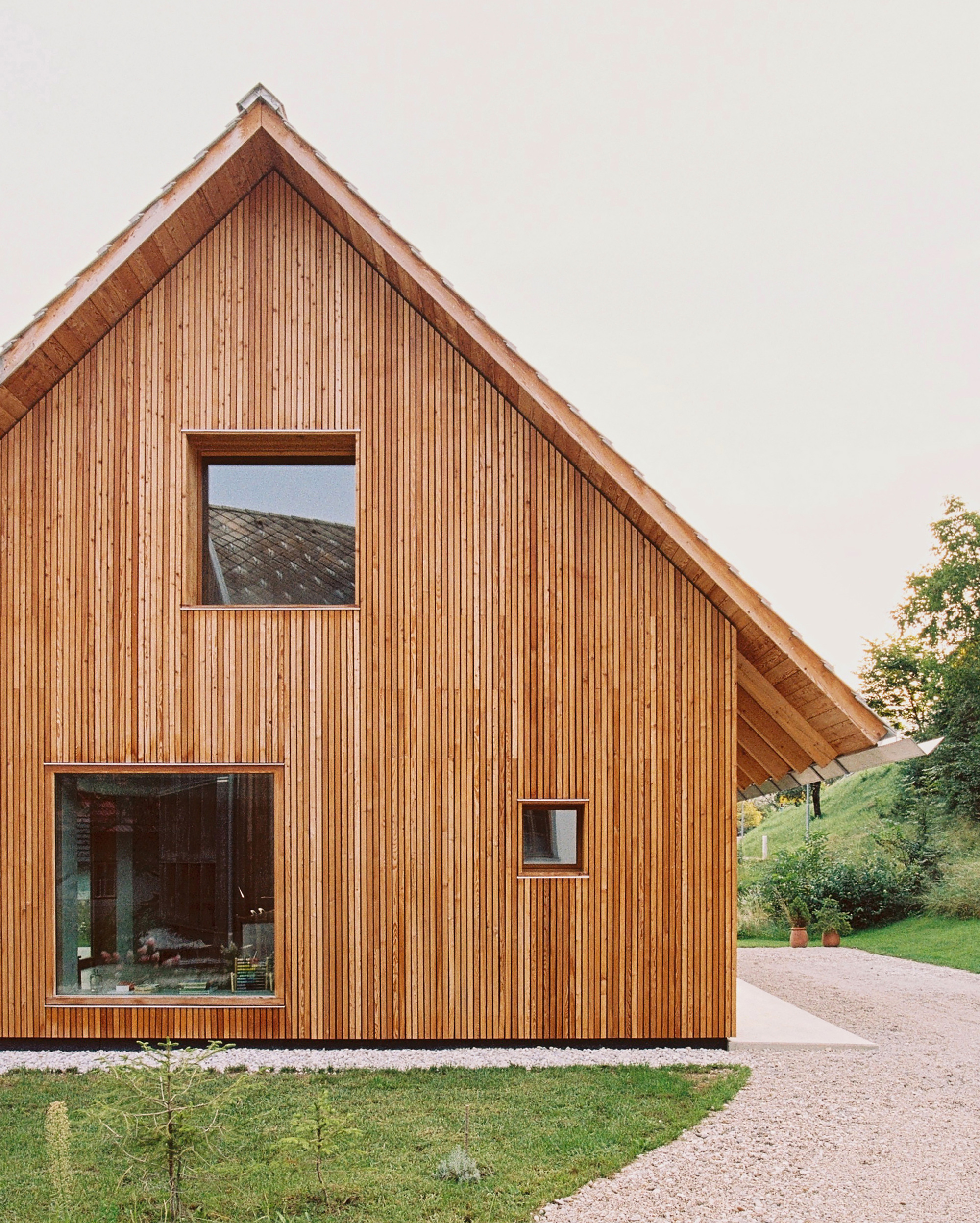Vertical timber-clad gable facade with square windows flanked by a gravel drive and lawn at dusk