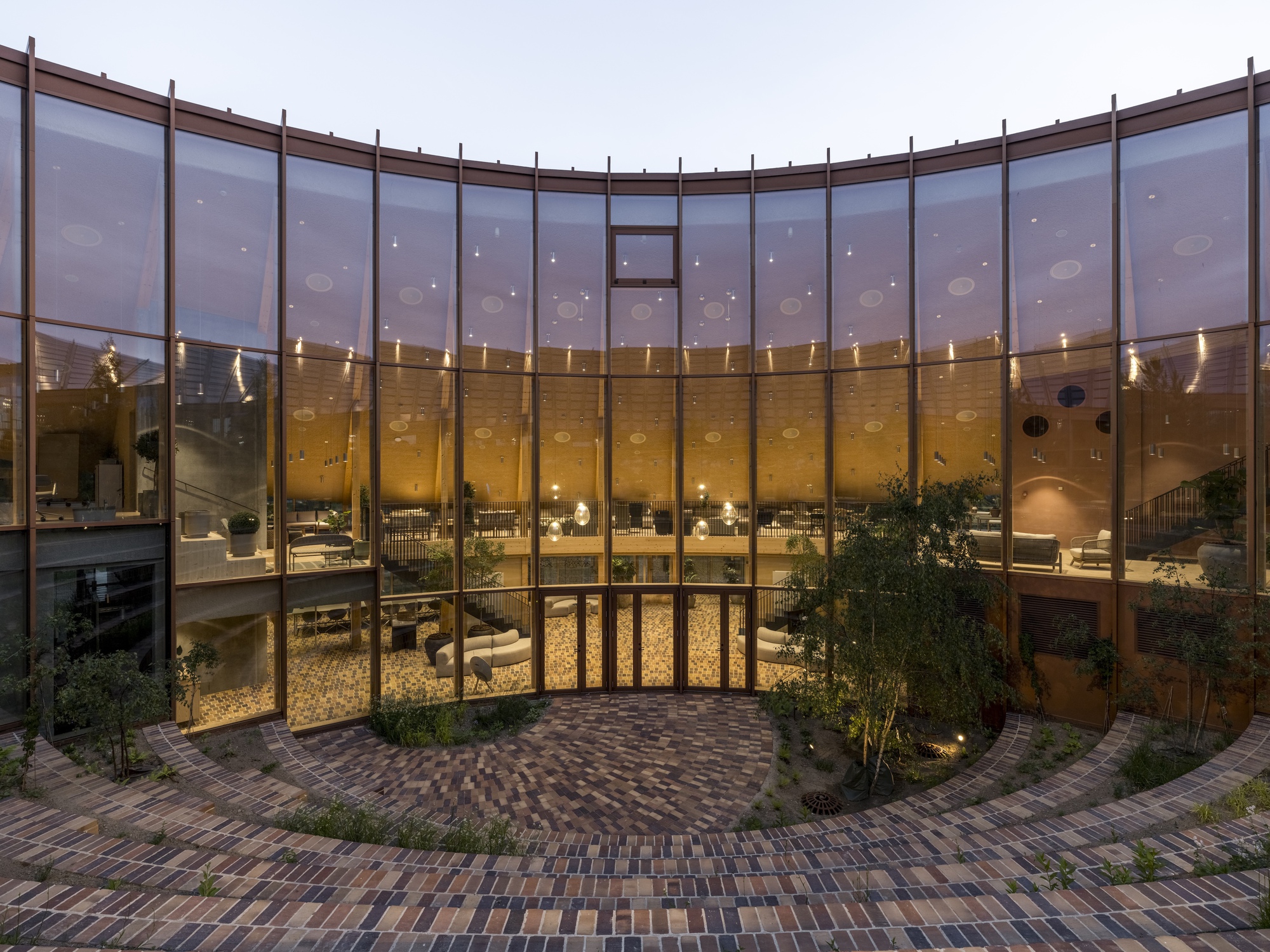 Interior courtyard with curved glass facade and brick paving at twilight with interior lighting visible