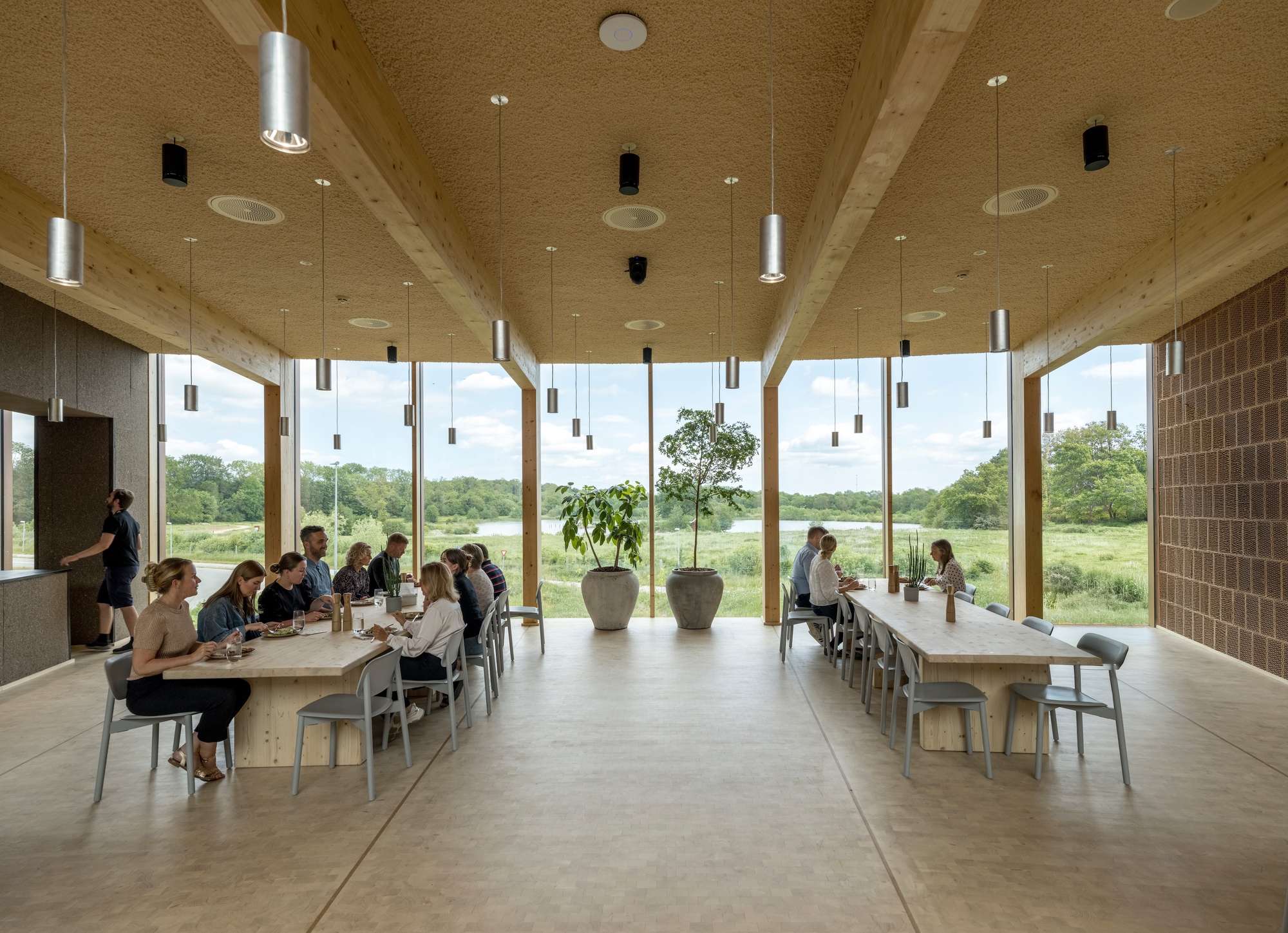 Interior dining space with long timber tables and floor-to-ceiling glazing framing a view of open meadow