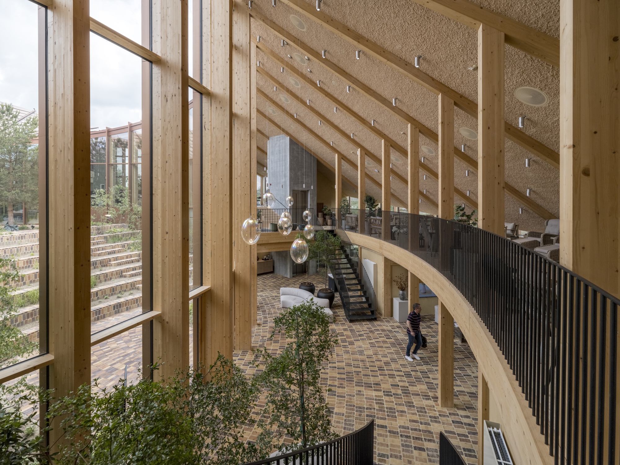 Upper-level view down to the brick-paved entrance lobby with sloped timber columns and planted trees
