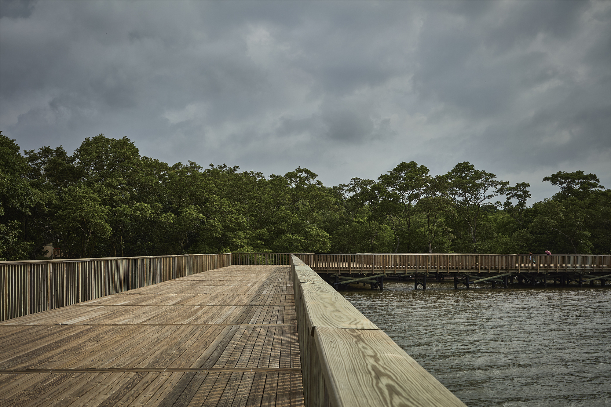 Wide timber boardwalk with vertical slat balustrades stretching across water toward tree line under grey clouds