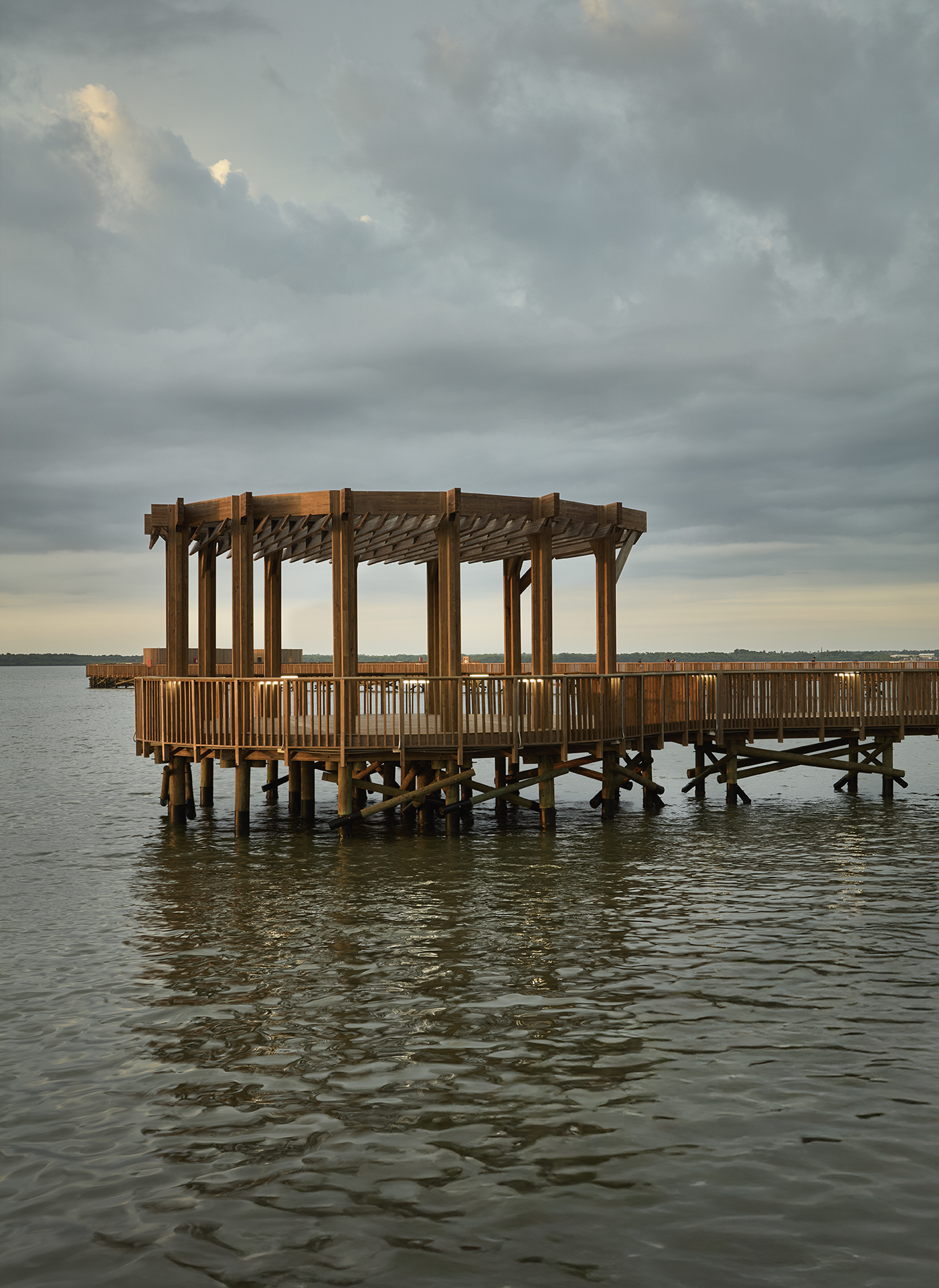 Timber pavilion with open pergola roof and vertical slat railings on pile supports above water at dusk