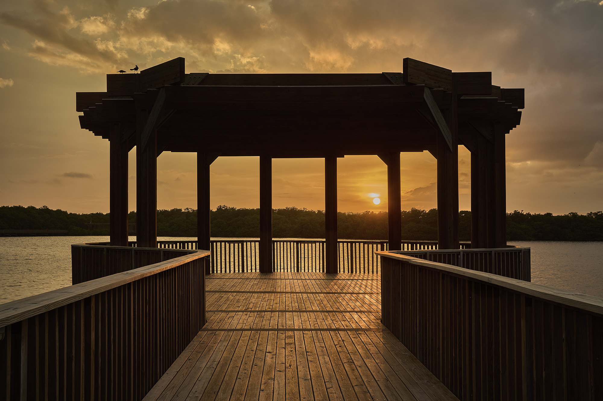 Timber deck pavilion with vertical slat railings and post-and-beam roof silhouetted at sunset over water