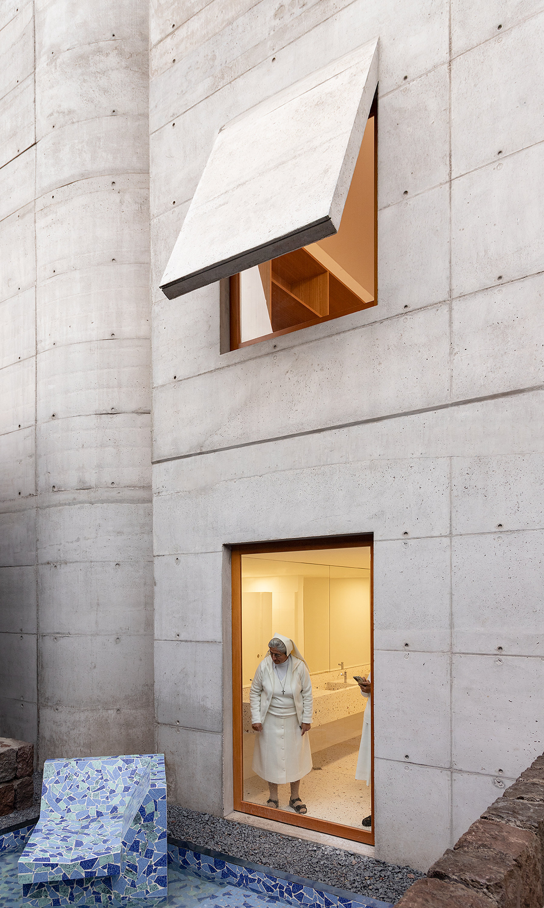 Board-formed concrete courtyard wall with two cantilevered window boxes and a person exiting at twilight