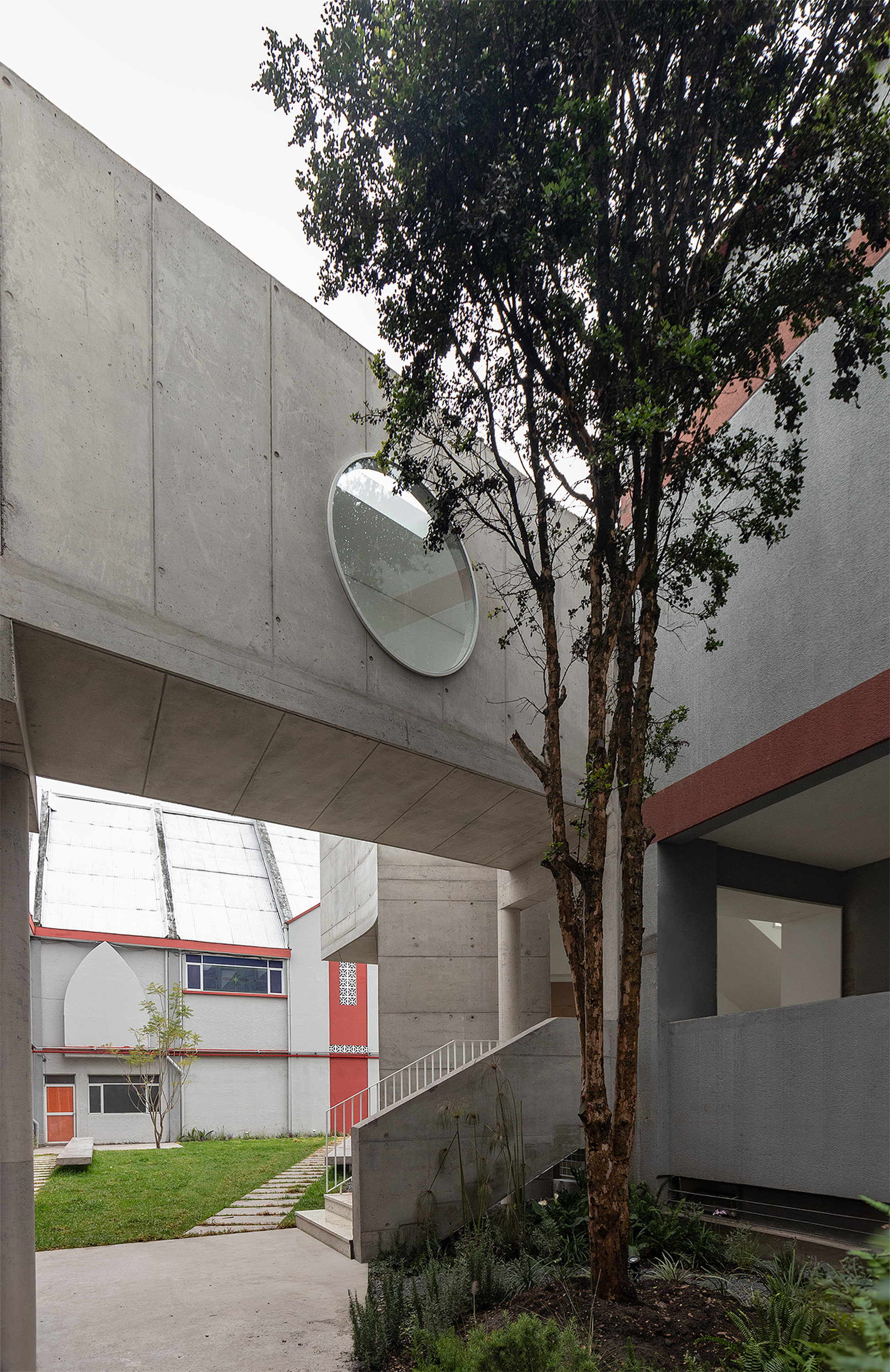 Courtyard view with concrete bridge overhead, circular mirror on the wall, and tree growing between volumes