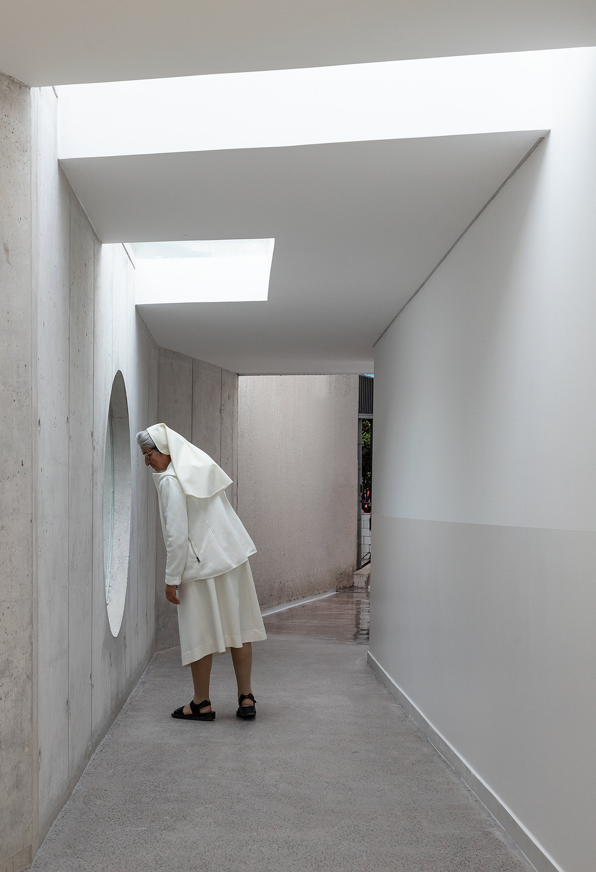 Skylit corridor with exposed concrete walls and a person standing beneath layered ceiling planes