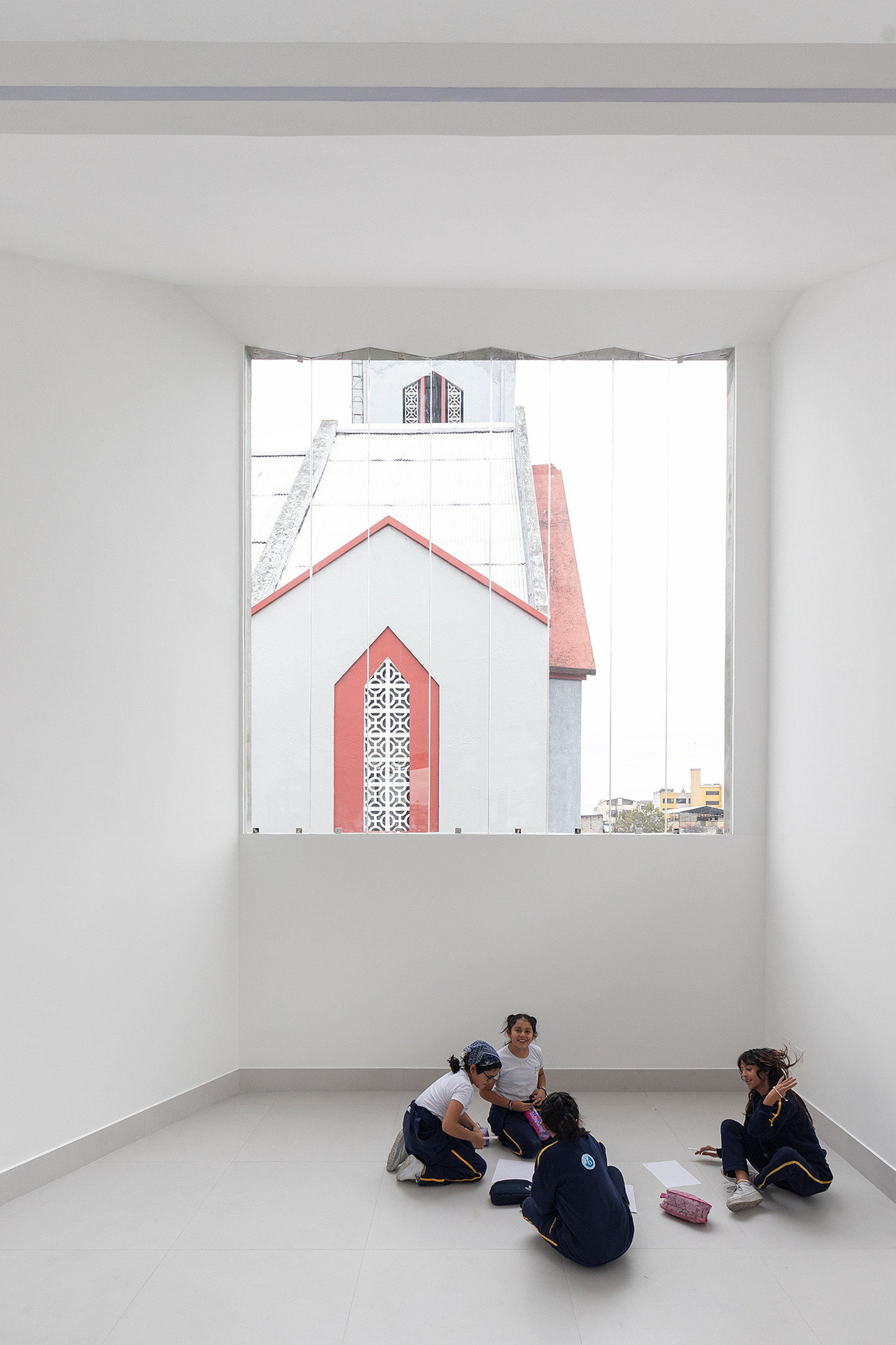 White interior room with students seated on the floor beneath a large window framing a church spire