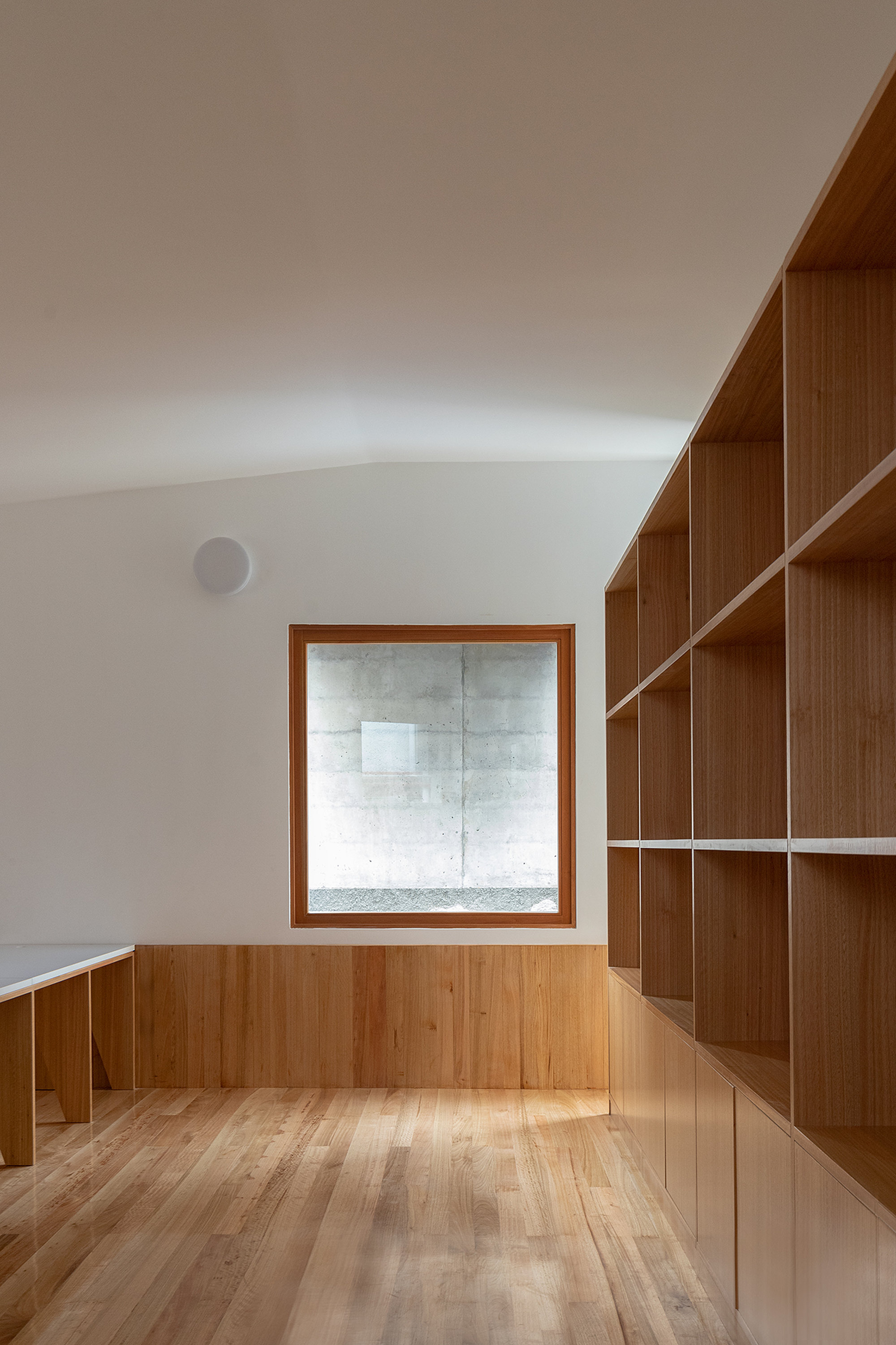 Library space with timber shelving and wainscoting surrounding a square window onto concrete exterior