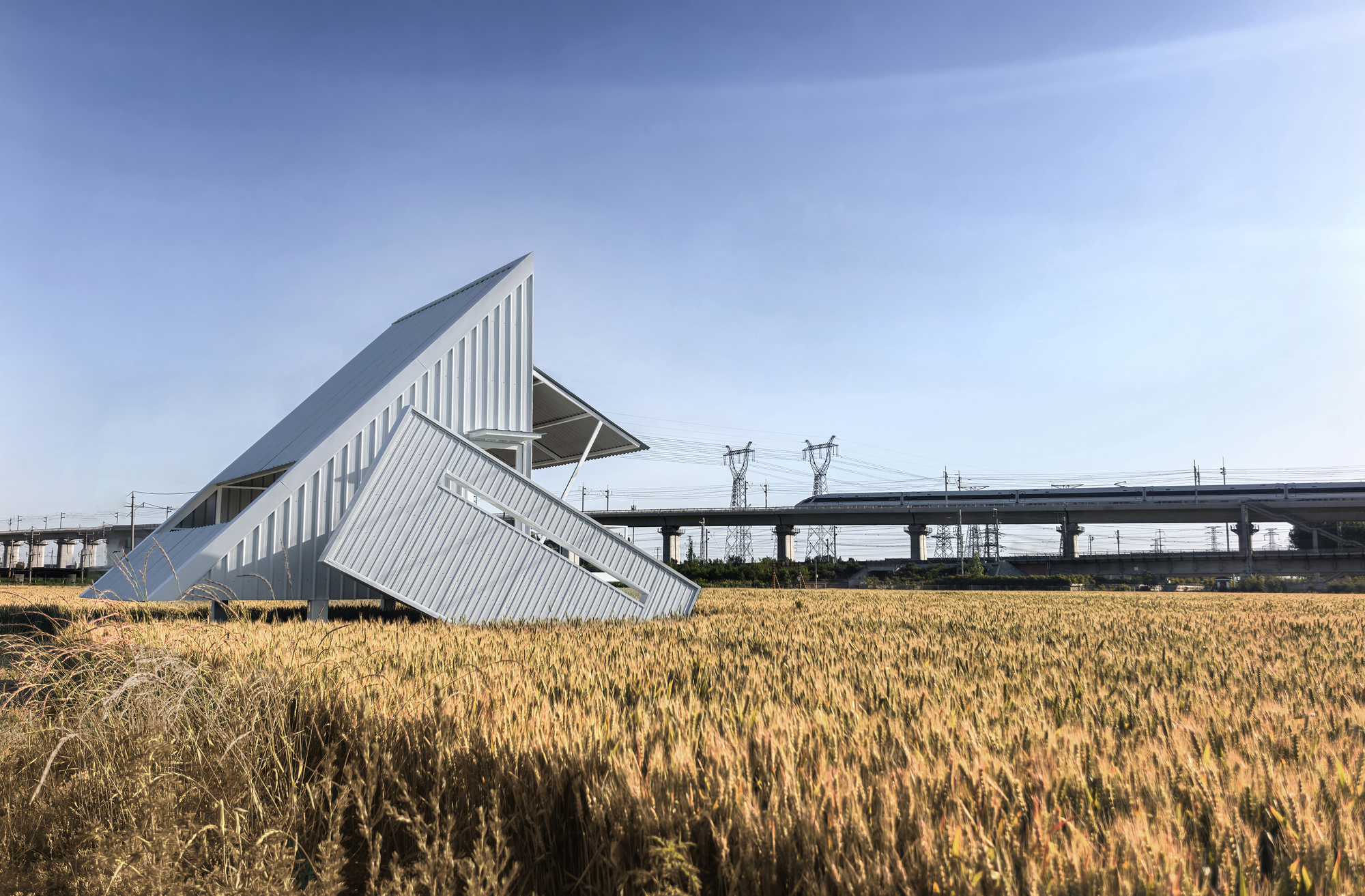 Corrugated metal pavilion with tilted volumes set in a dry wheat field beside overhead viaduct