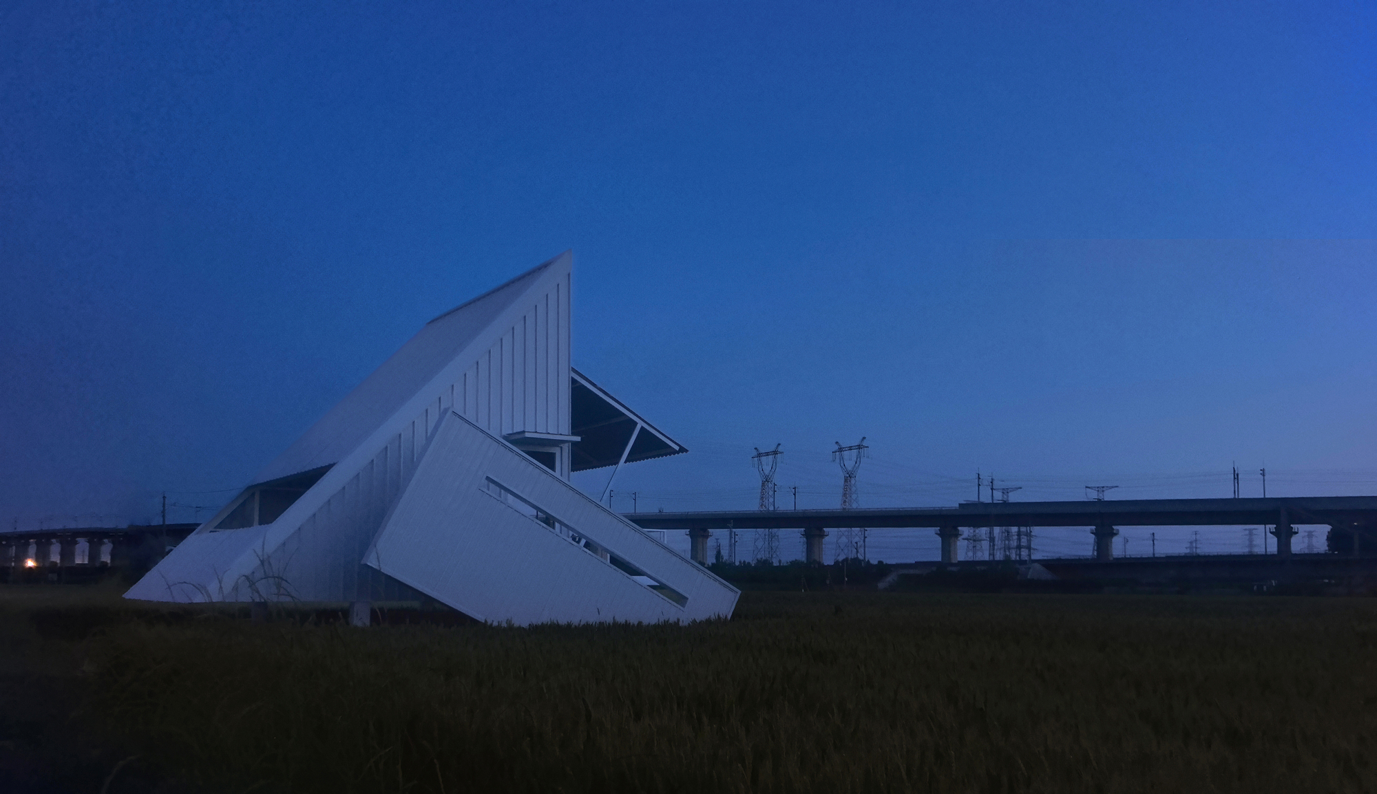 Folded metal pavilion in grassland at dusk with elevated highway spanning the background