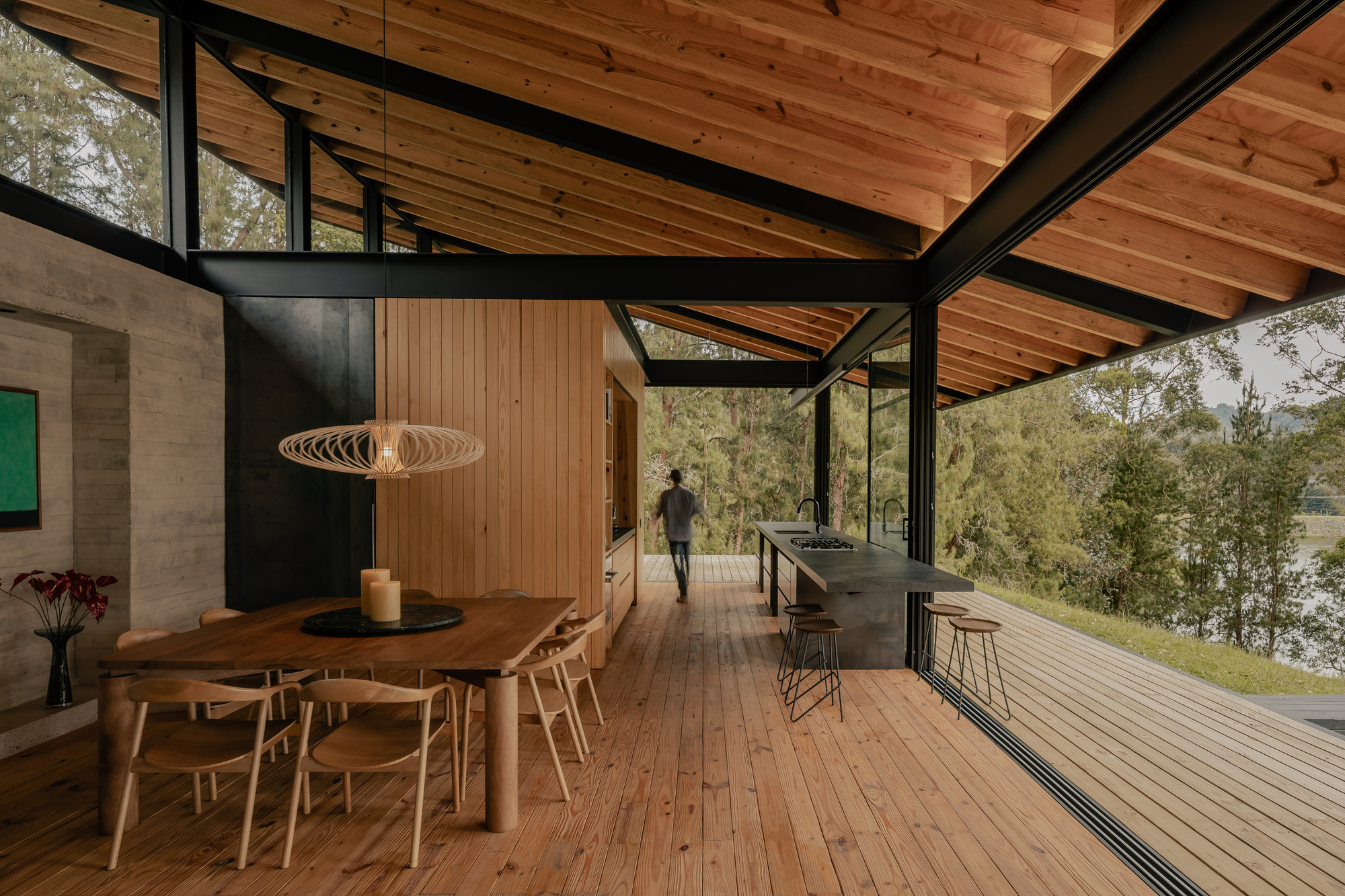 Open-plan dining and kitchen space with timber ceiling, black steel structure, and figure walking toward the landscape