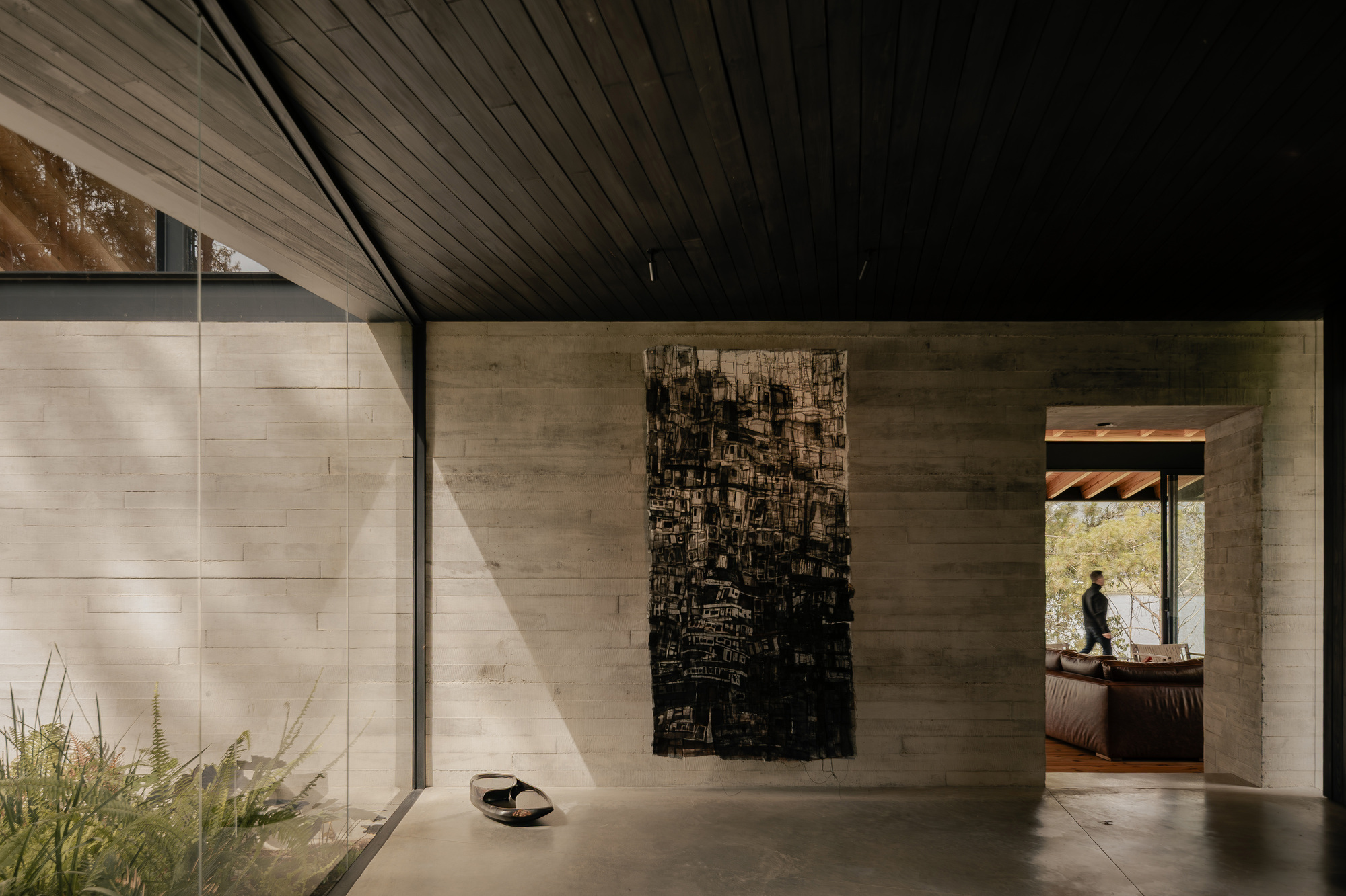 Gallery hallway with board-formed concrete walls, dark timber ceiling, and figure silhouetted in distant doorway