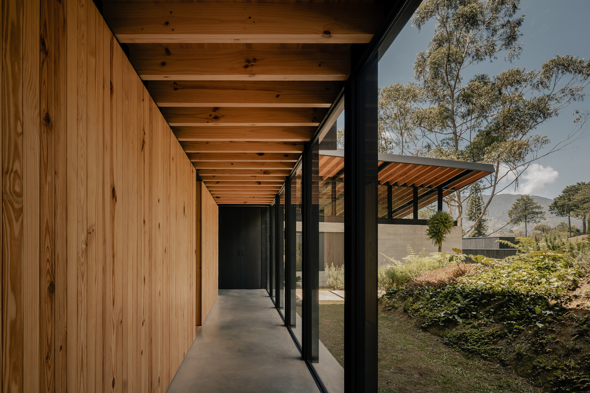 Covered walkway with timber ceiling beams and vertical wood wall alongside planted garden under cloudy sky