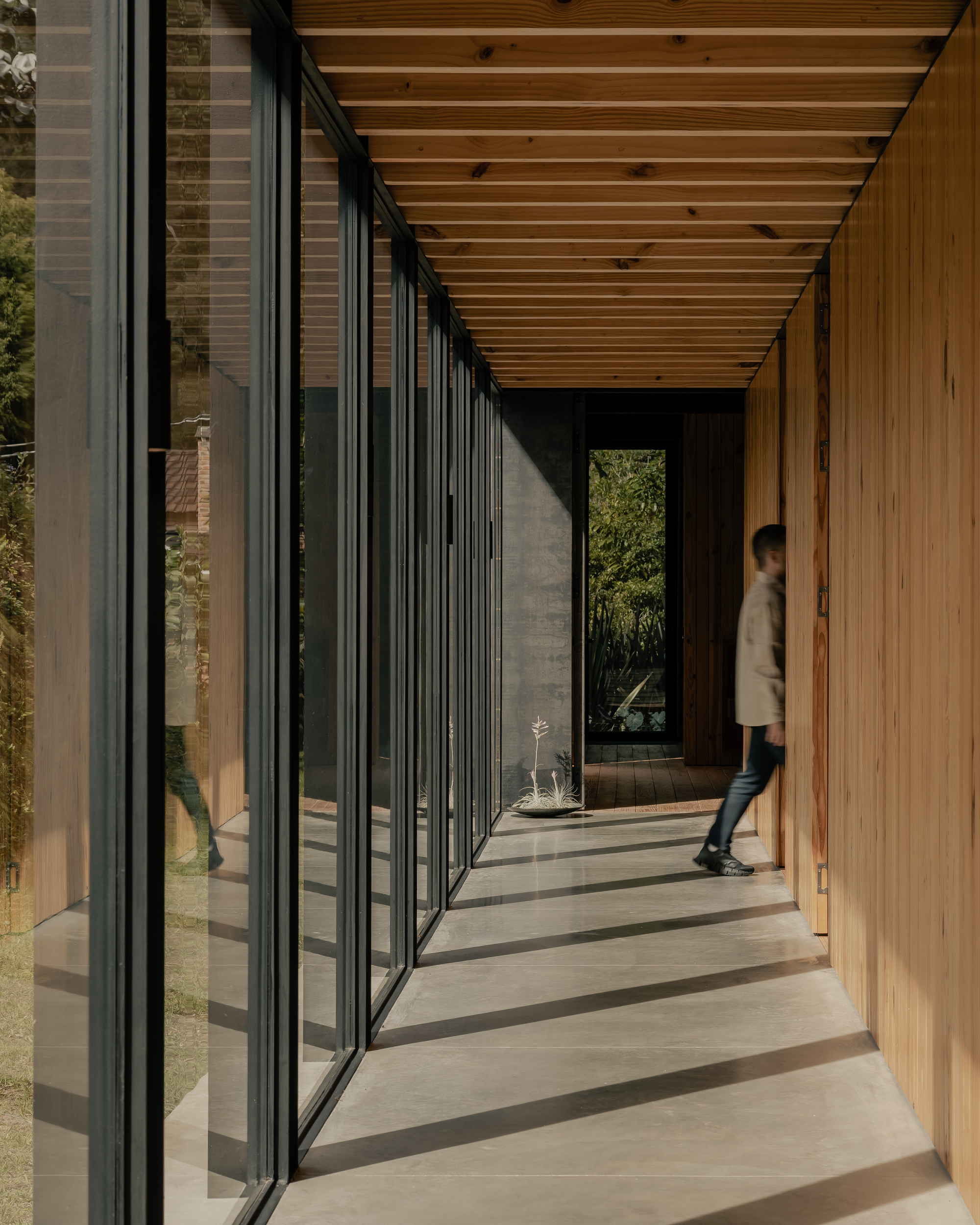 Glazed corridor with person walking on concrete floor beneath exposed timber joists casting shadows