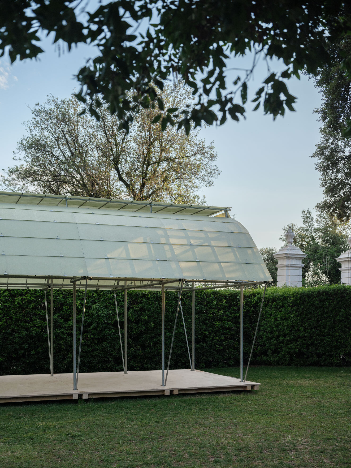 Translucent pavilion roof on slender steel columns set in a garden framed by overhanging tree branches