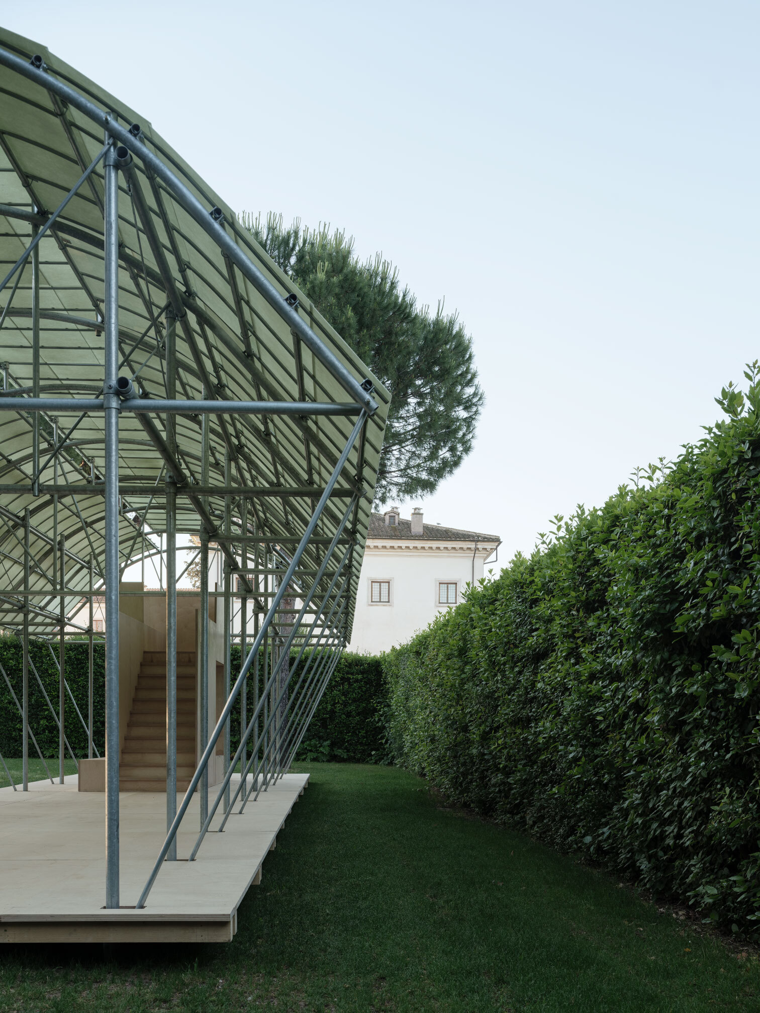 Close-up of the exposed triangulated steel frame supporting the translucent panel roof against hedges