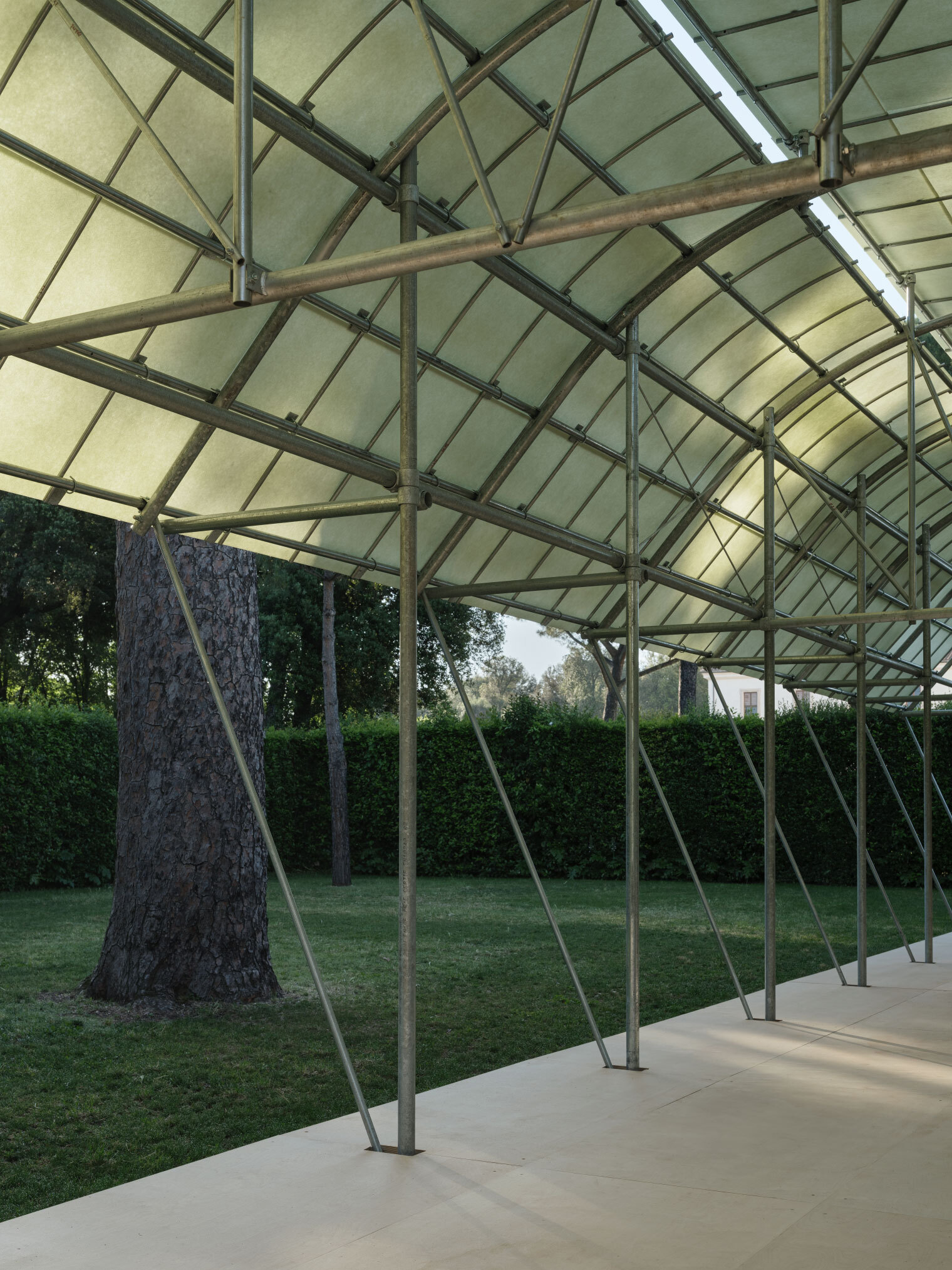 Covered pavilion with translucent fabric roof and steel frame supported by diagonal columns in a grassy clearing