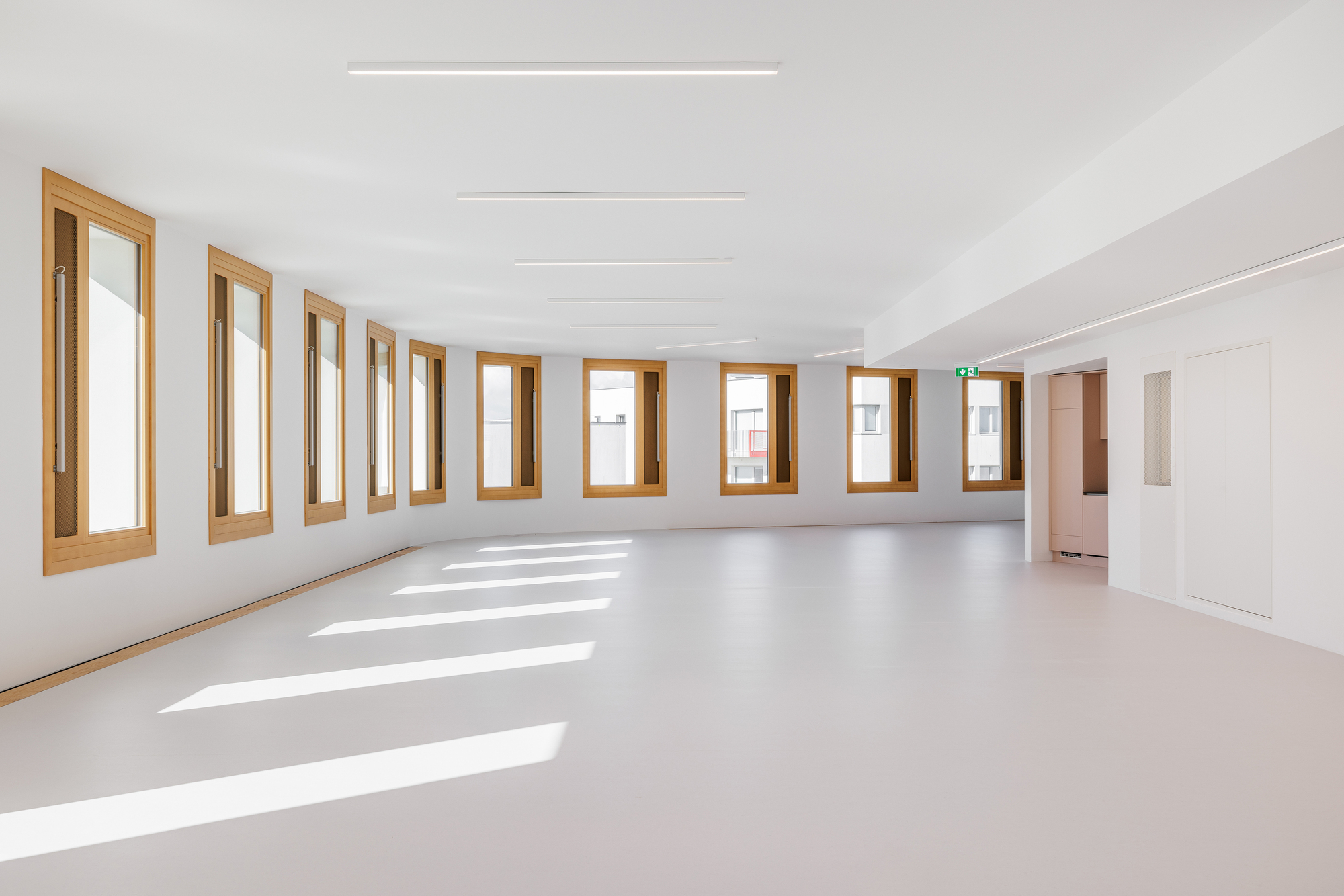 Empty white interior with timber-framed windows casting sunlight across the pale floor