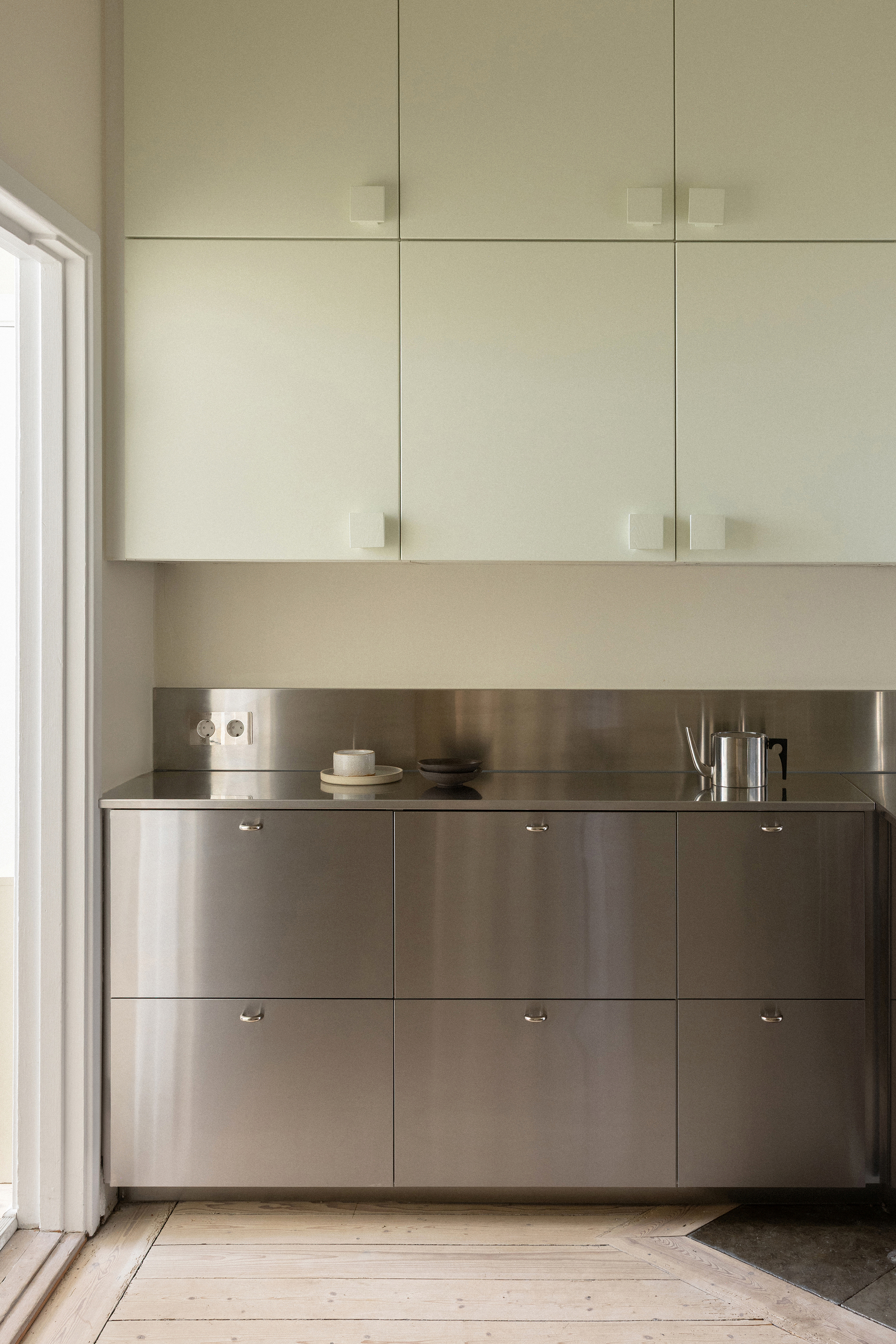 Kitchen counter with brushed stainless steel base cabinets below pale upper cabinetry and a window